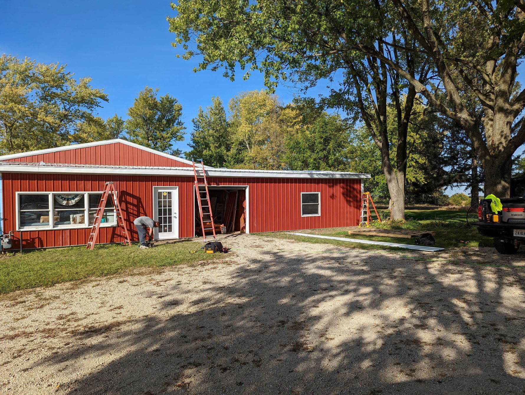 Red metal building with workers, ladders, and gravel driveway under a blue sky and trees.