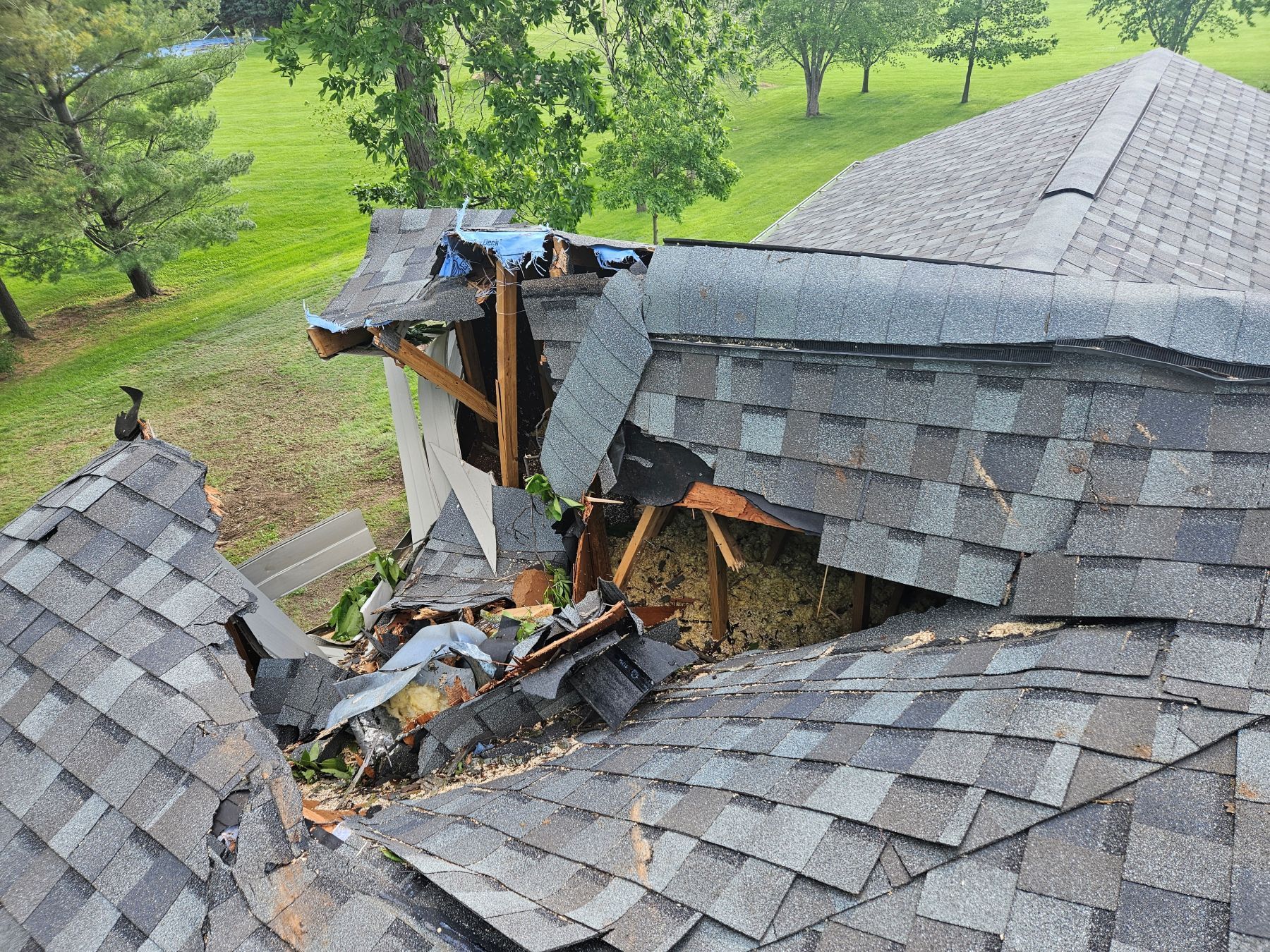 Damaged roof with missing shingles and exposed wooden structure, in a grassy setting.