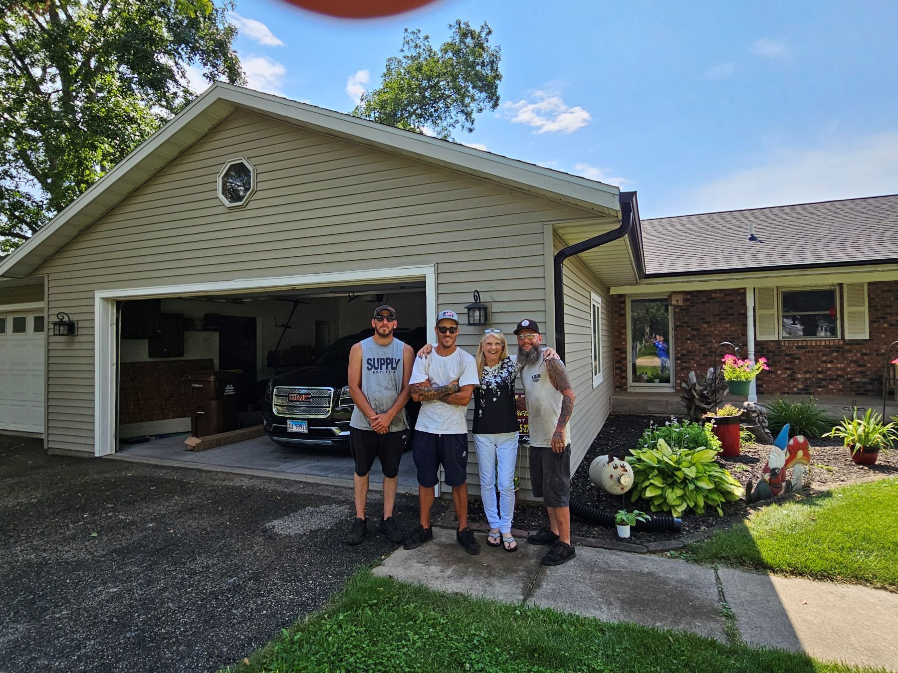 Four people stand in front of a house. The house has a garage. Sunny day.