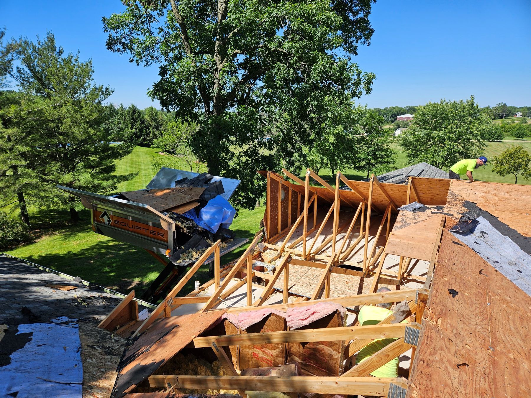 Roof under construction: Shingles removed, wooden supports exposed, worker in yellow shirt. Outdoors on a sunny day.