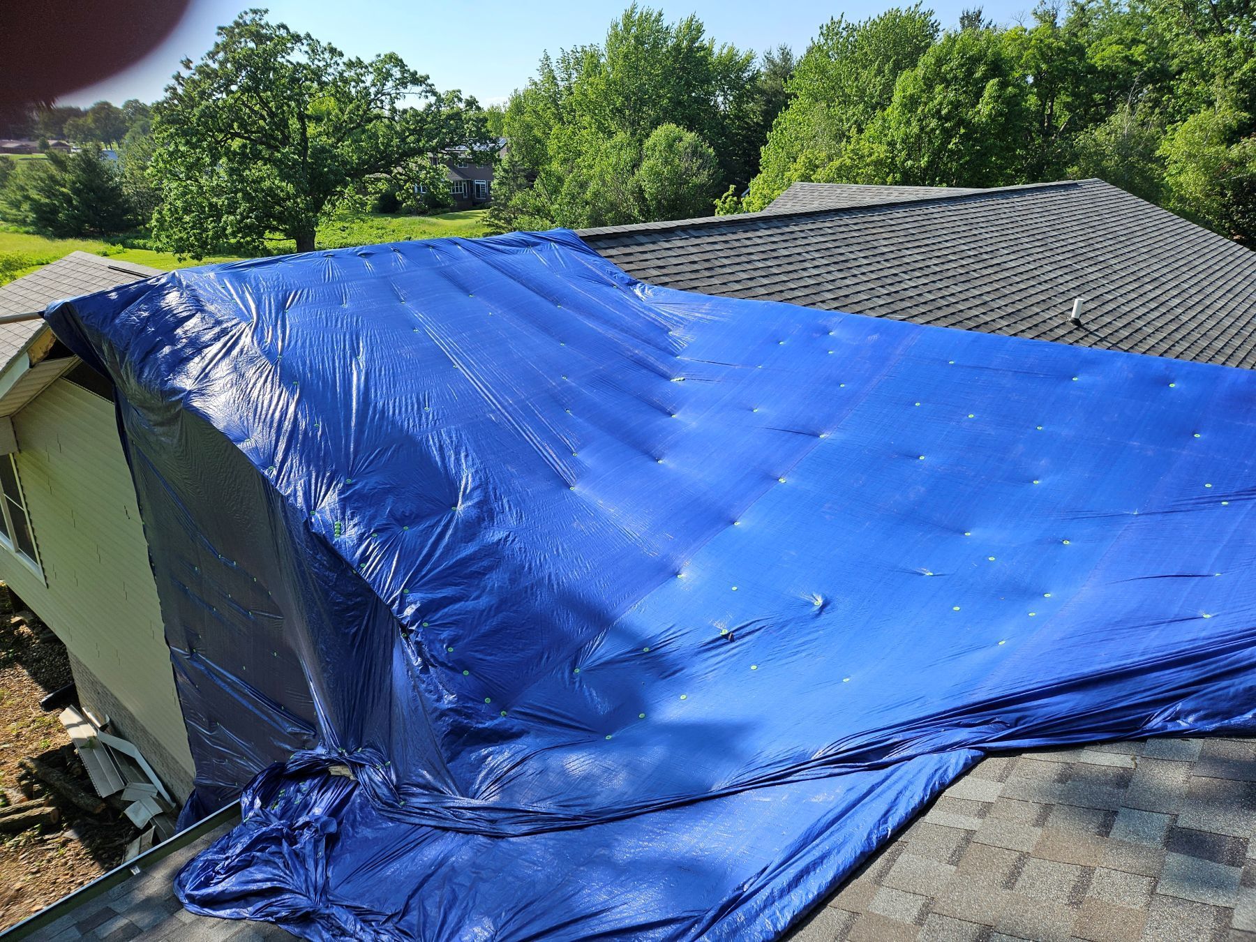 Blue tarp covering a damaged roof on a house, trees in the background.
