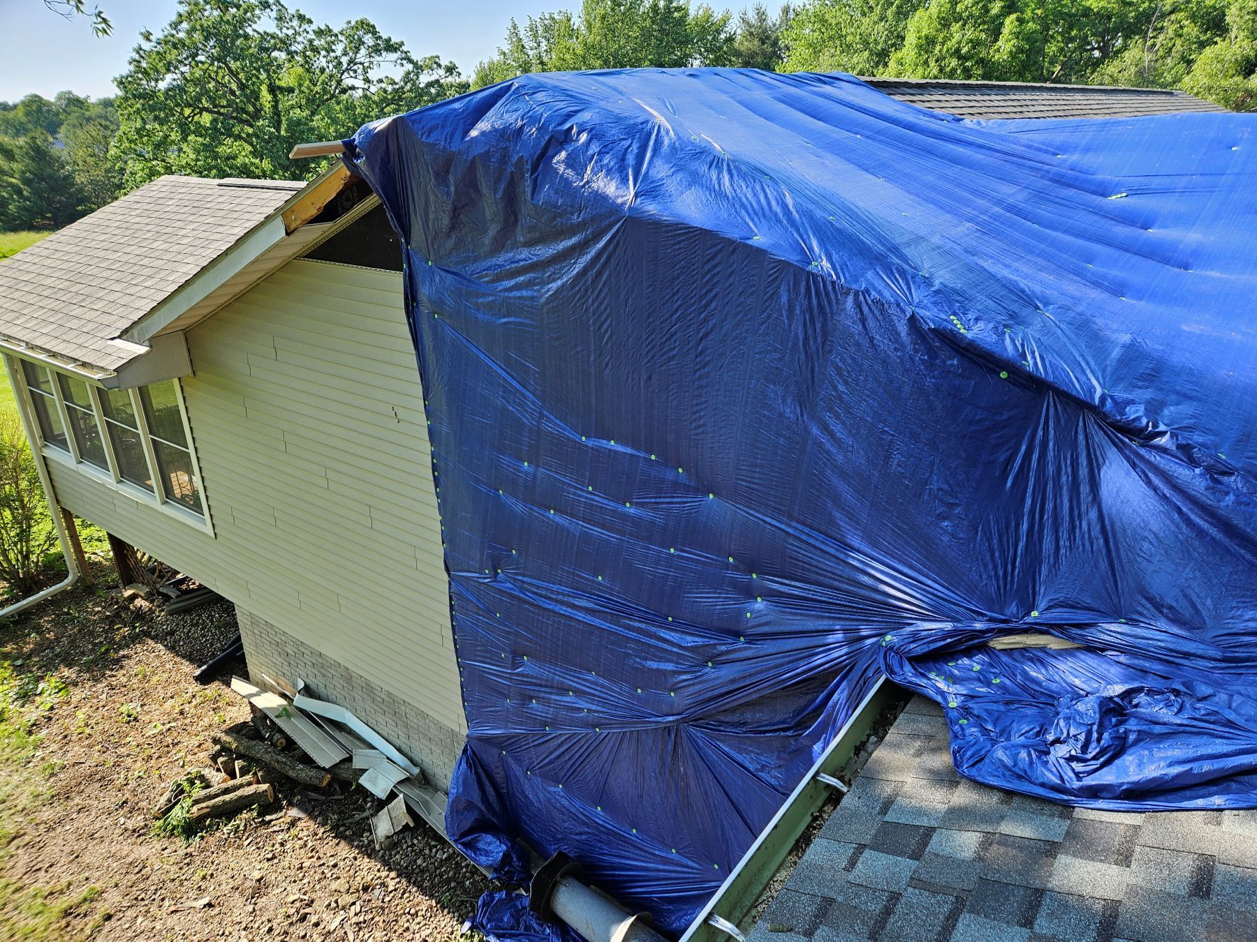 A house with a blue tarp covering a damaged roof. Green trees and grass are in the background.