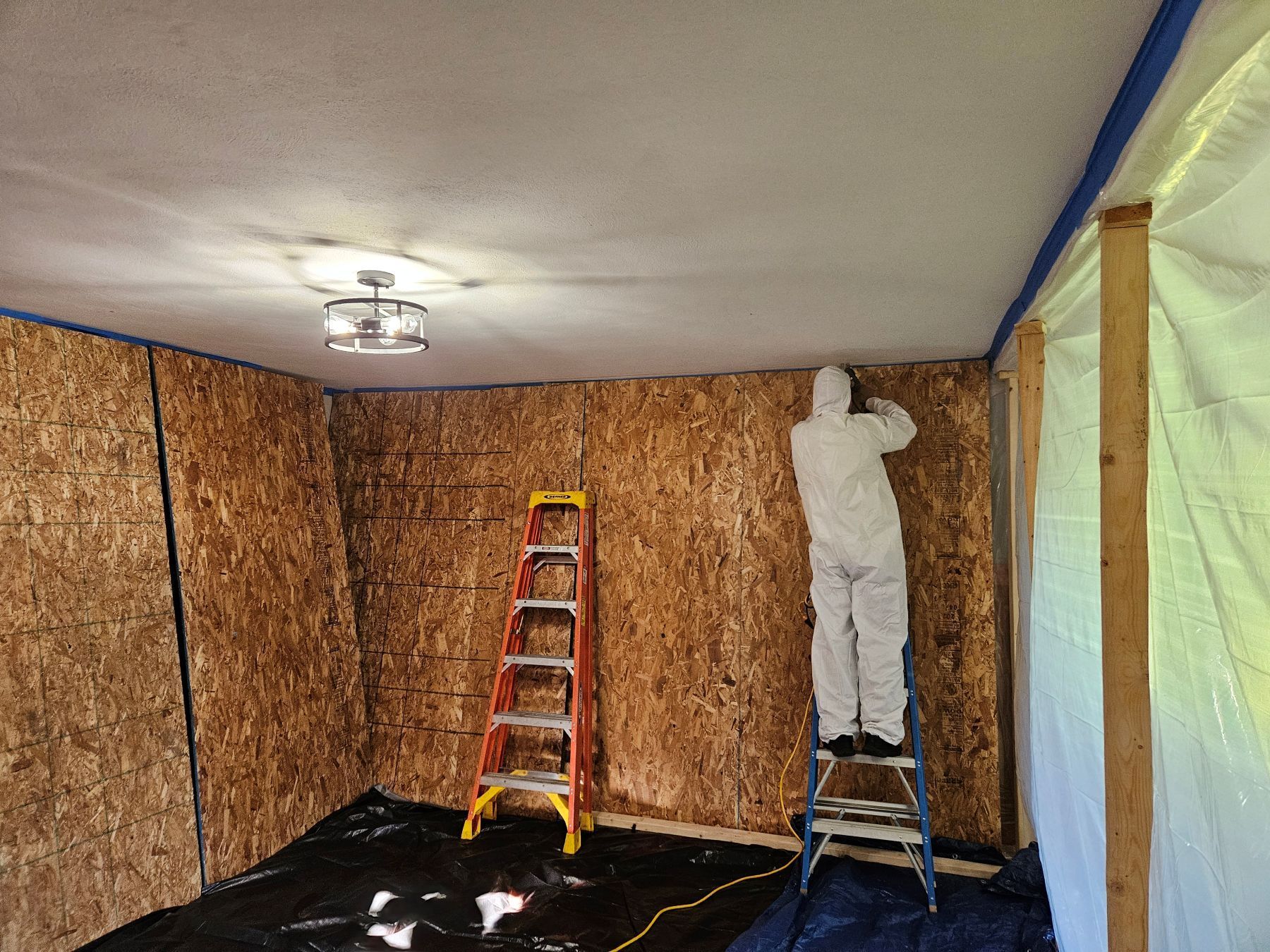 Person in protective suit on ladder, working in room with exposed wood walls, tarp, and ladder.