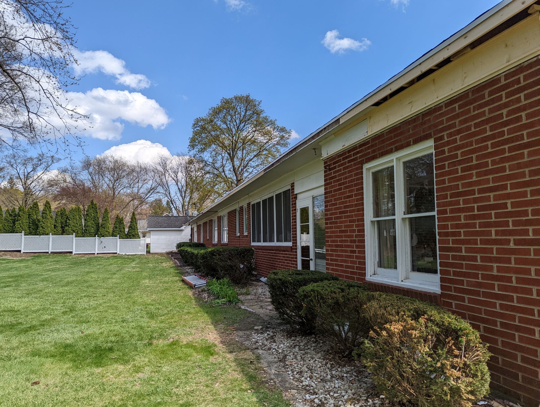 Brick ranch-style house with green lawn, bushes, and white fence under a partly cloudy blue sky.