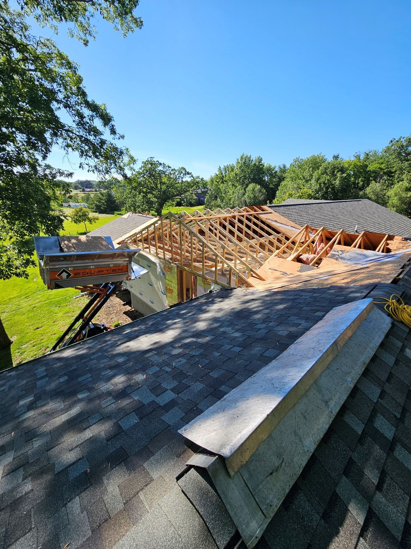 Roof construction in progress; wooden framework visible, partially completed. Blue sky, green trees in background.
