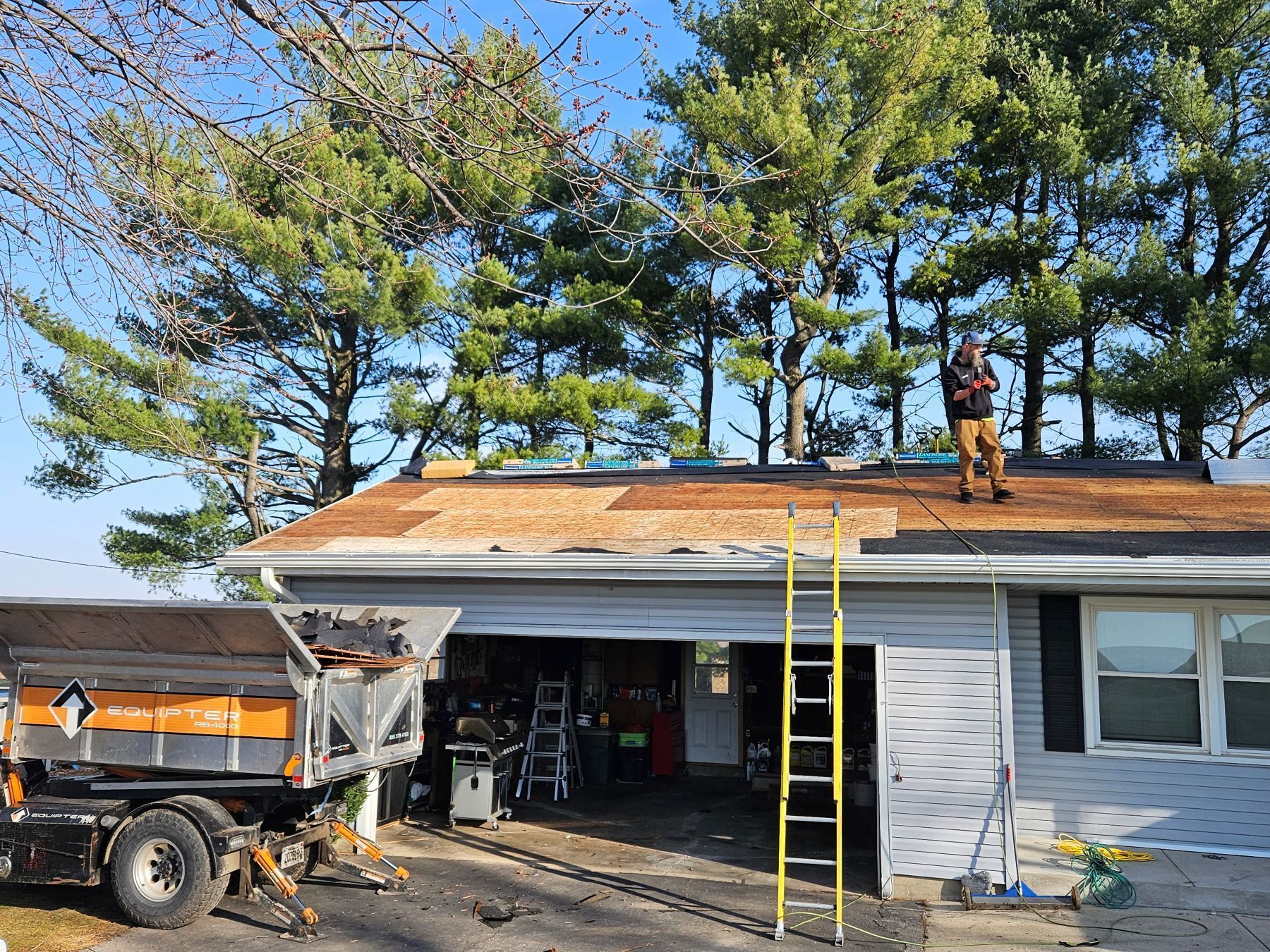 Roofer on a roof, removing old shingles with a truck parked beside a garage. Sunny day.