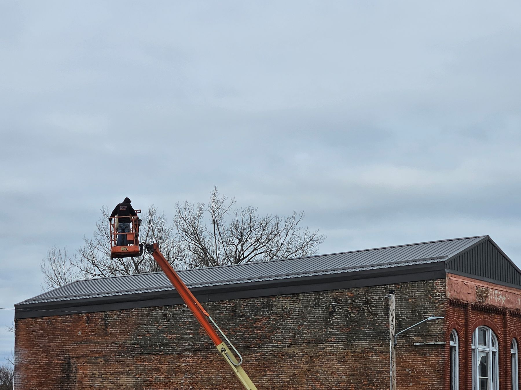 Person in a lift working on a brick building's roof under an overcast sky.