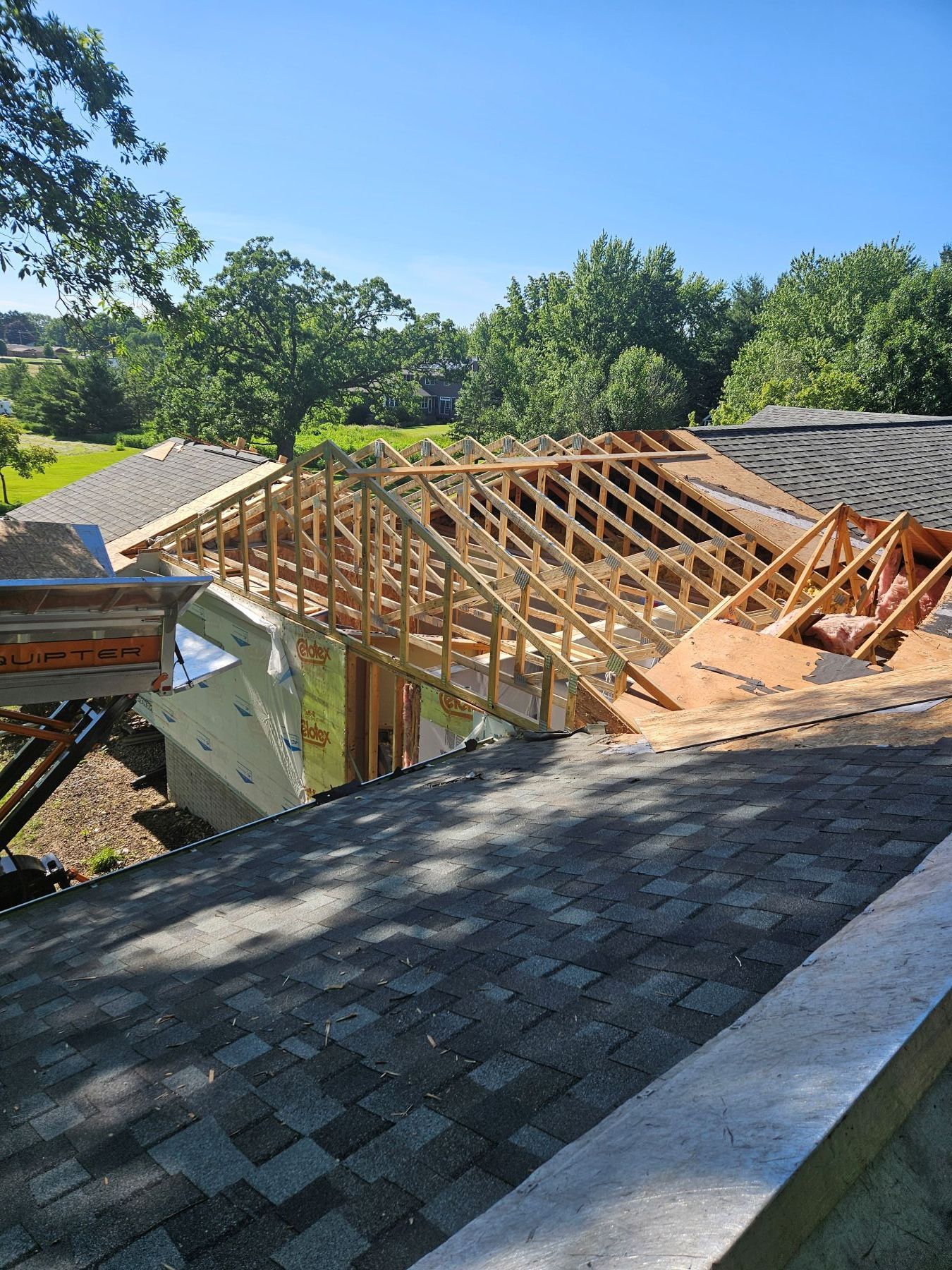 Construction site: New wooden roof frame being built on a house with existing asphalt shingle roof. Sunny day.