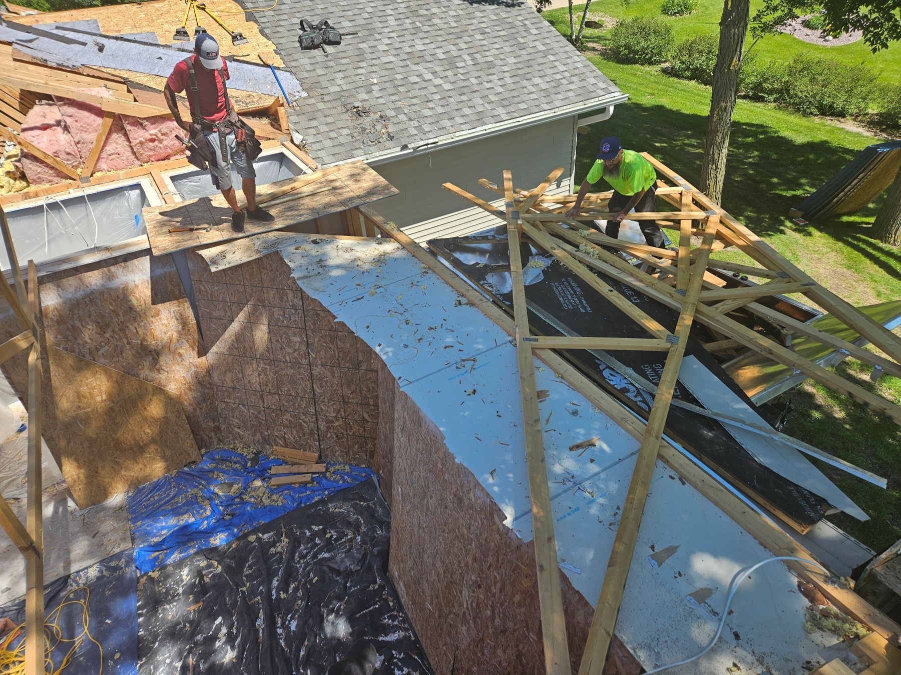 Two construction workers building a roof on a sunny day. One is on the roof, the other on a wooden frame.