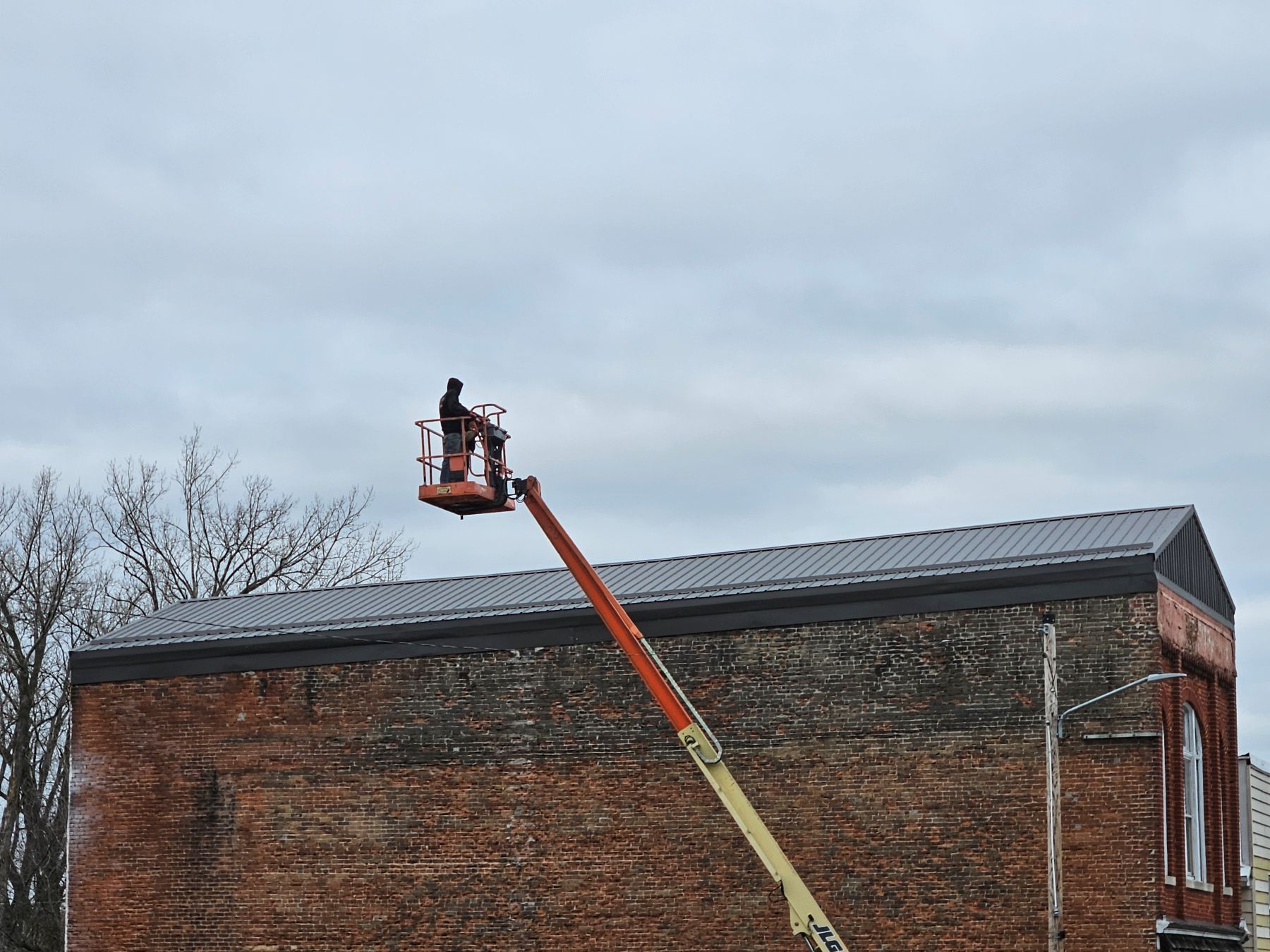 Person on lift working on a brick building with a dark roof under a cloudy sky.