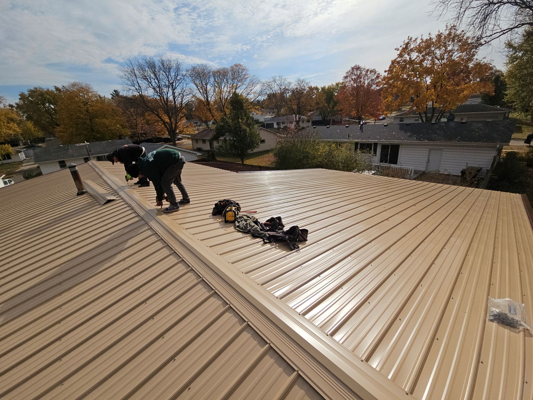 Two people installing tan metal roofing on a house, sunny day, trees in background.