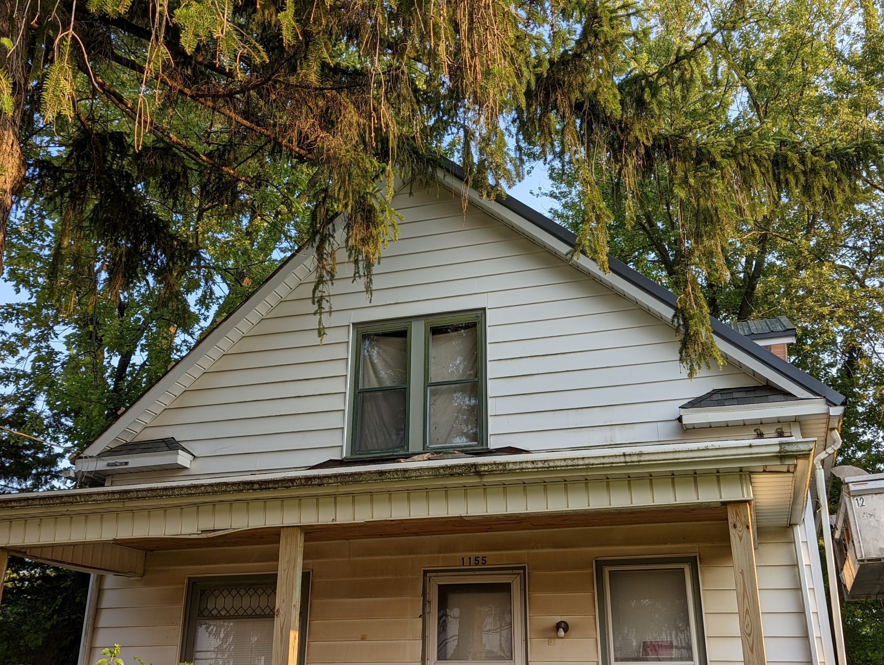 Two-story house with white siding and a covered porch. Some windows are boarded up. Trees in the background.