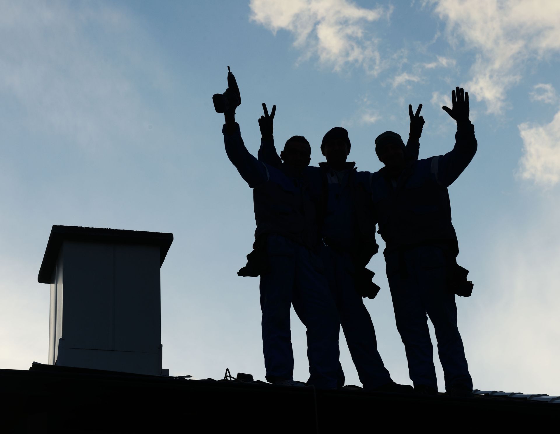 Silhouetted figures on a rooftop celebrate, raising arms and tool. Blue sky background.
