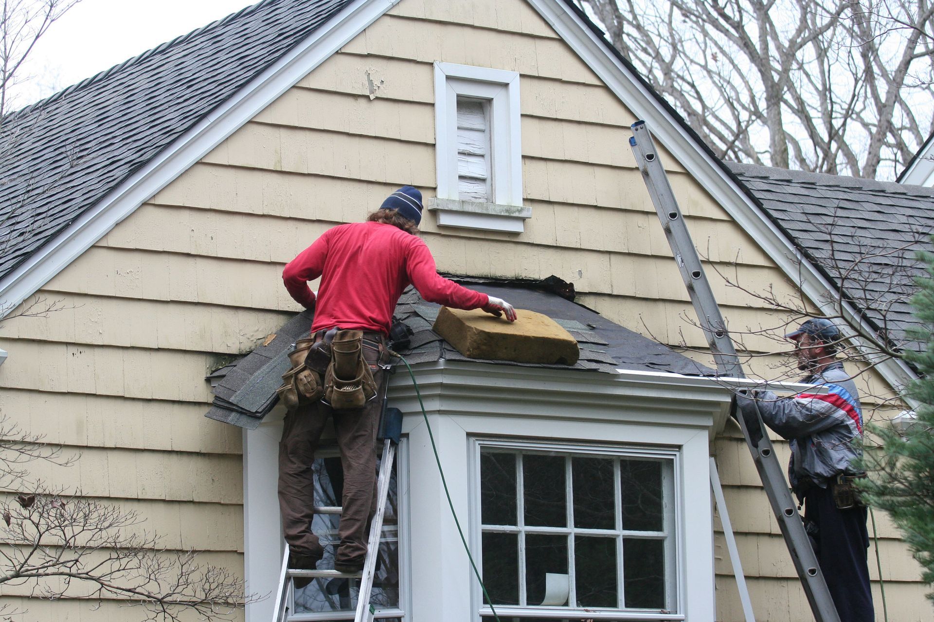 Two roofers on ladders repairing a gable roof. One holds insulation, the other looks on. Beige siding, cloudy sky.