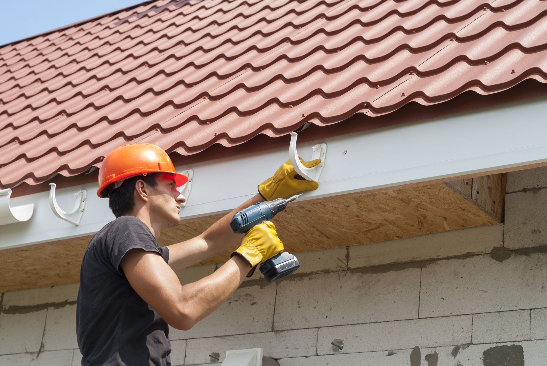 Construction worker in orange hard hat, installing a white gutter on a red tiled roof with a power drill.