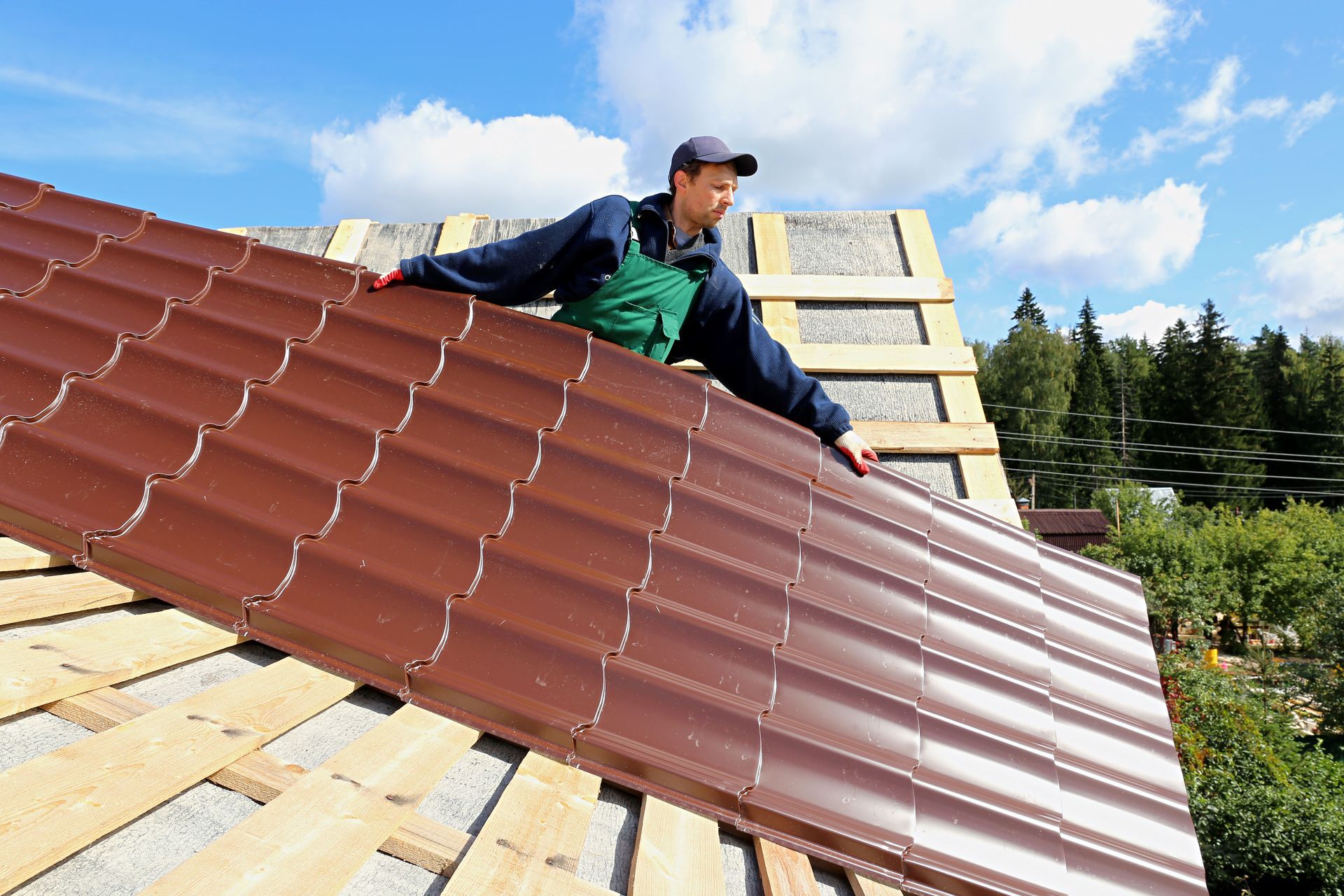 Roofer installs brown metal roofing on a house under construction; blue sky, sunny day.