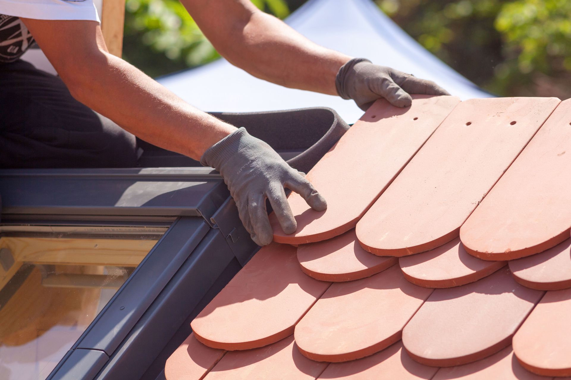 Person installing reddish-brown roof tiles on a skylight, wearing gloves.