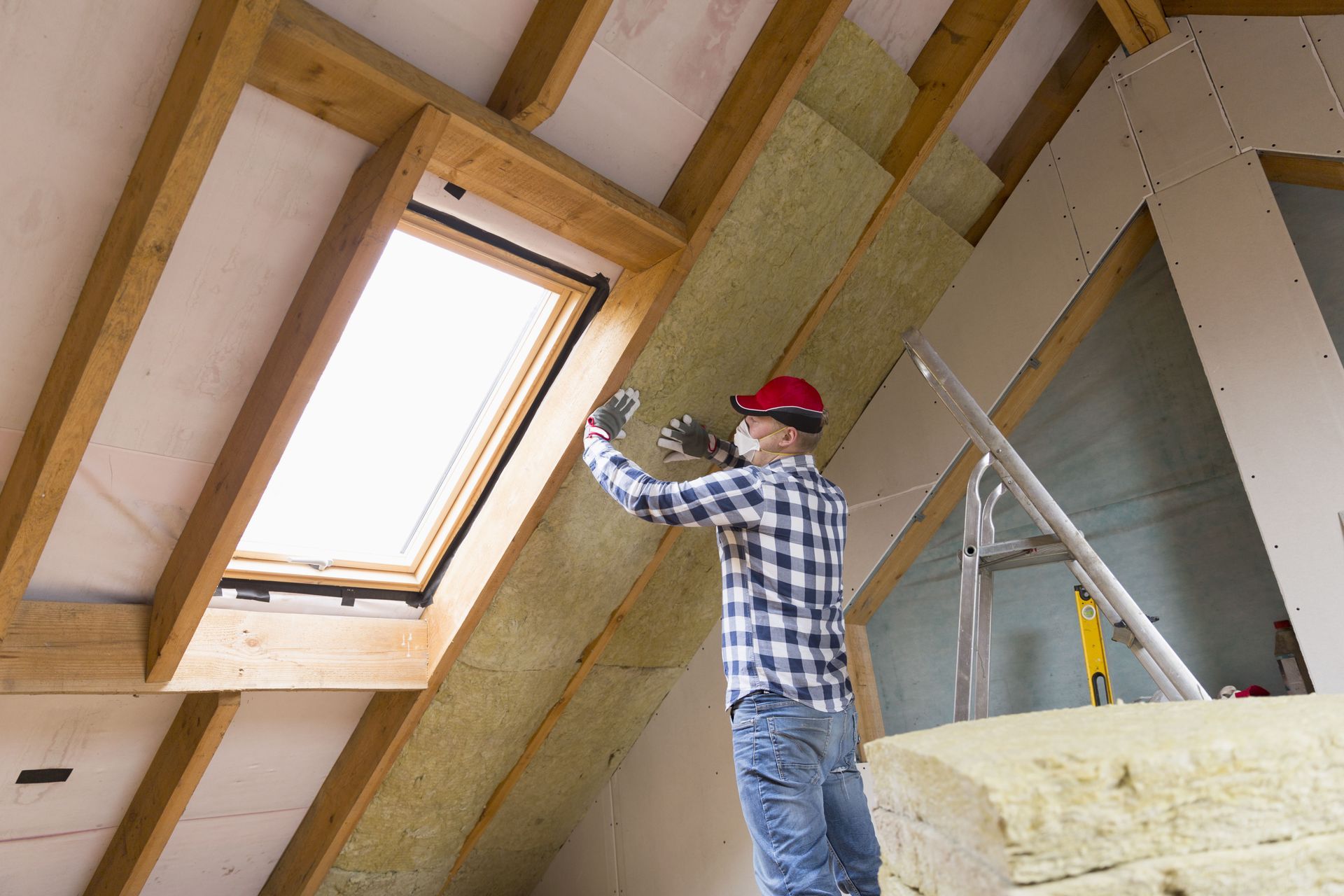 Person installing insulation in an attic. They are wearing a hard hat, working near a skylight.