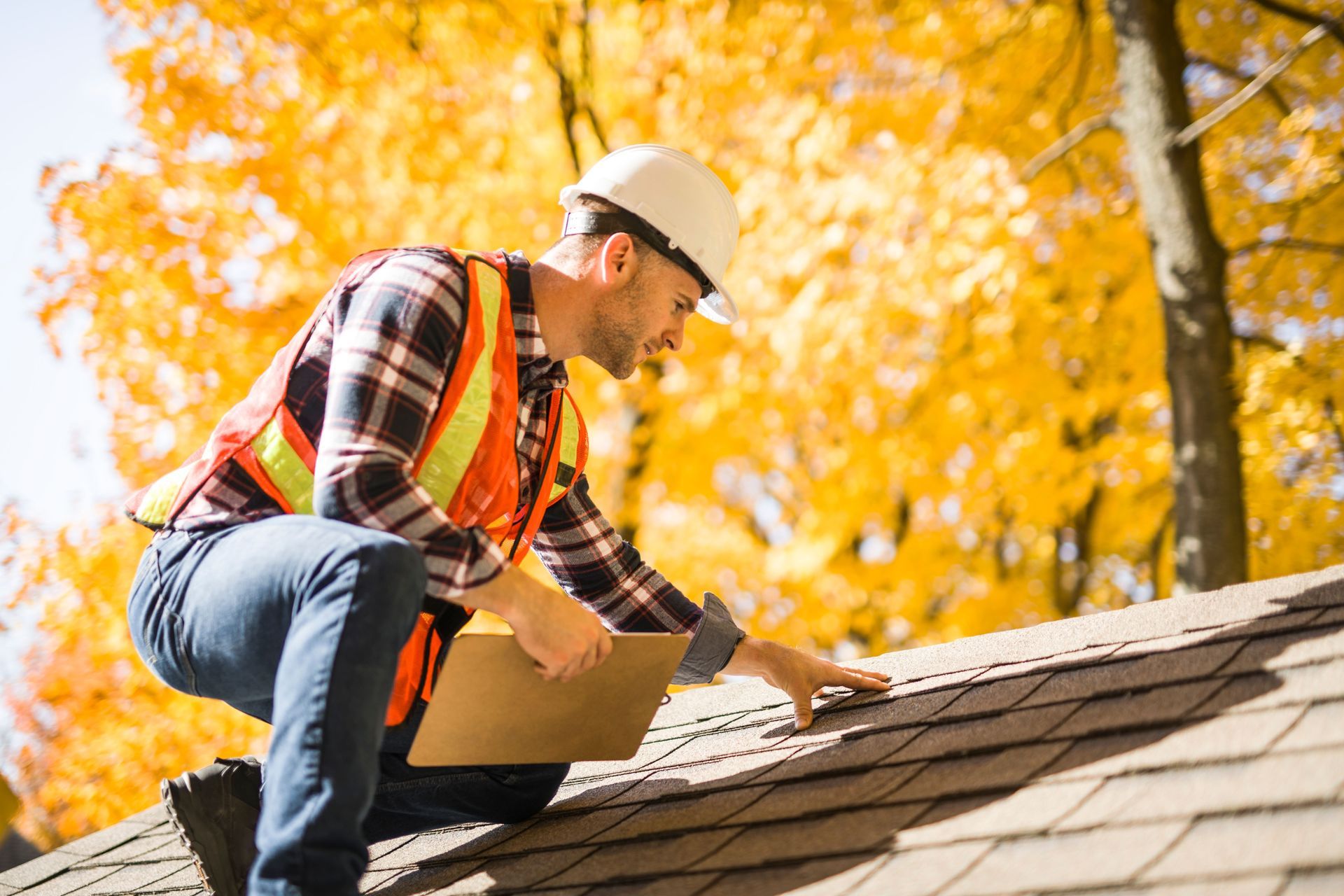 A construction worker in a hard hat and safety vest kneeling on a roof while inspecting shingles, with autumn trees behind.