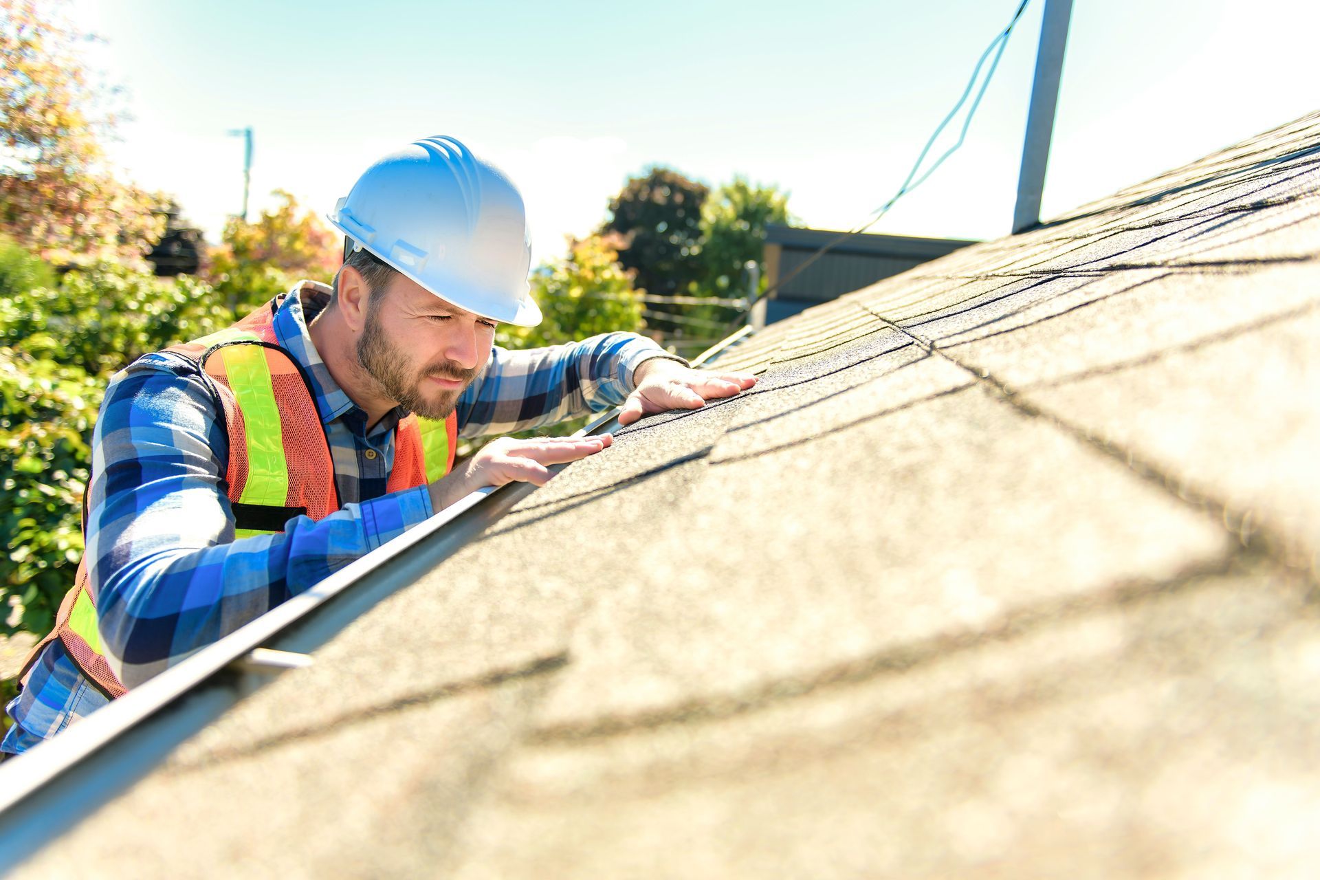 A worker in a high-visibility vest and hard hat inspects shingles on a residential roof.