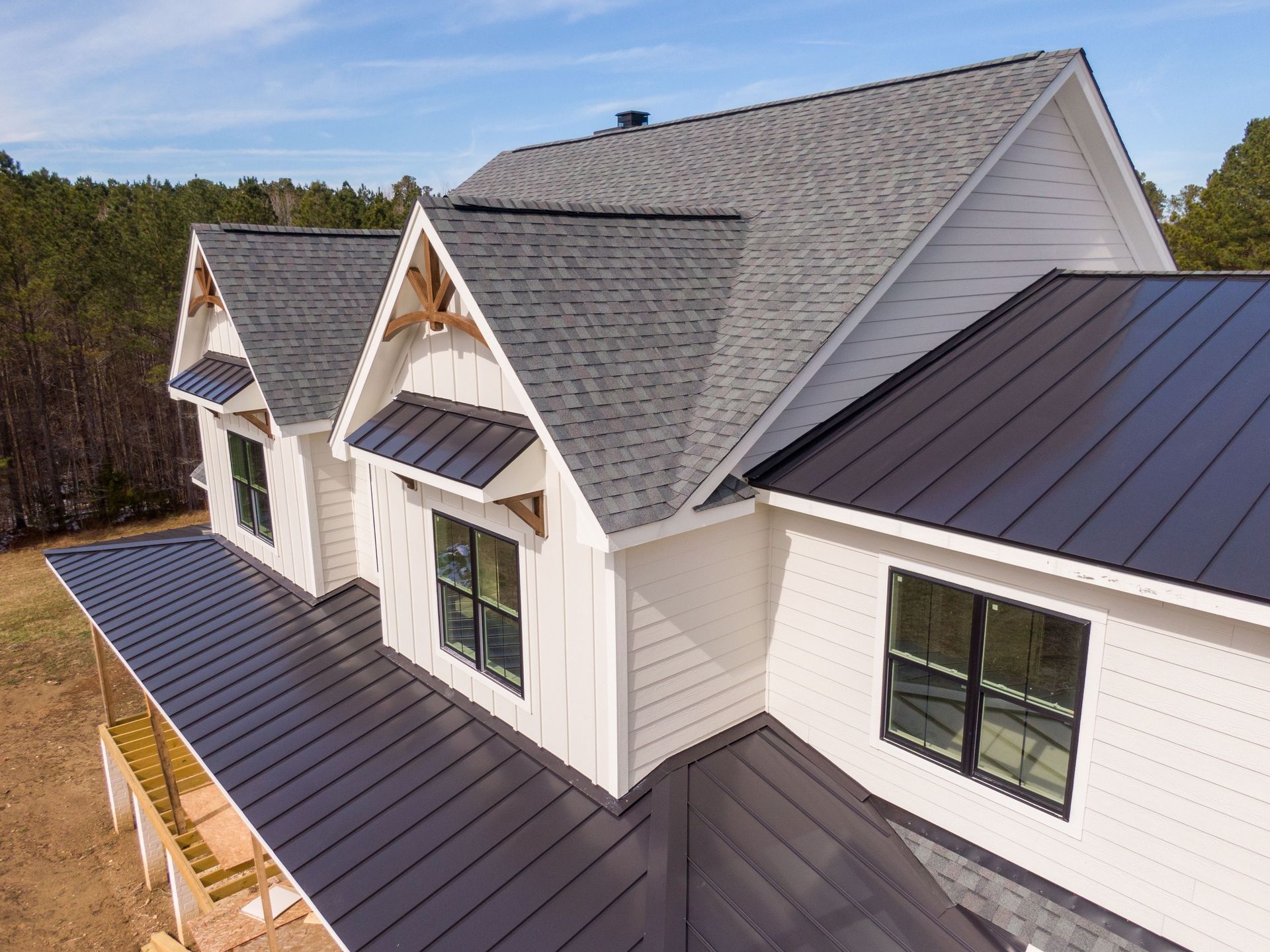 An aerial view of a modern farmhouse with a mix of dark metal and shingle roofing against a wooded background.