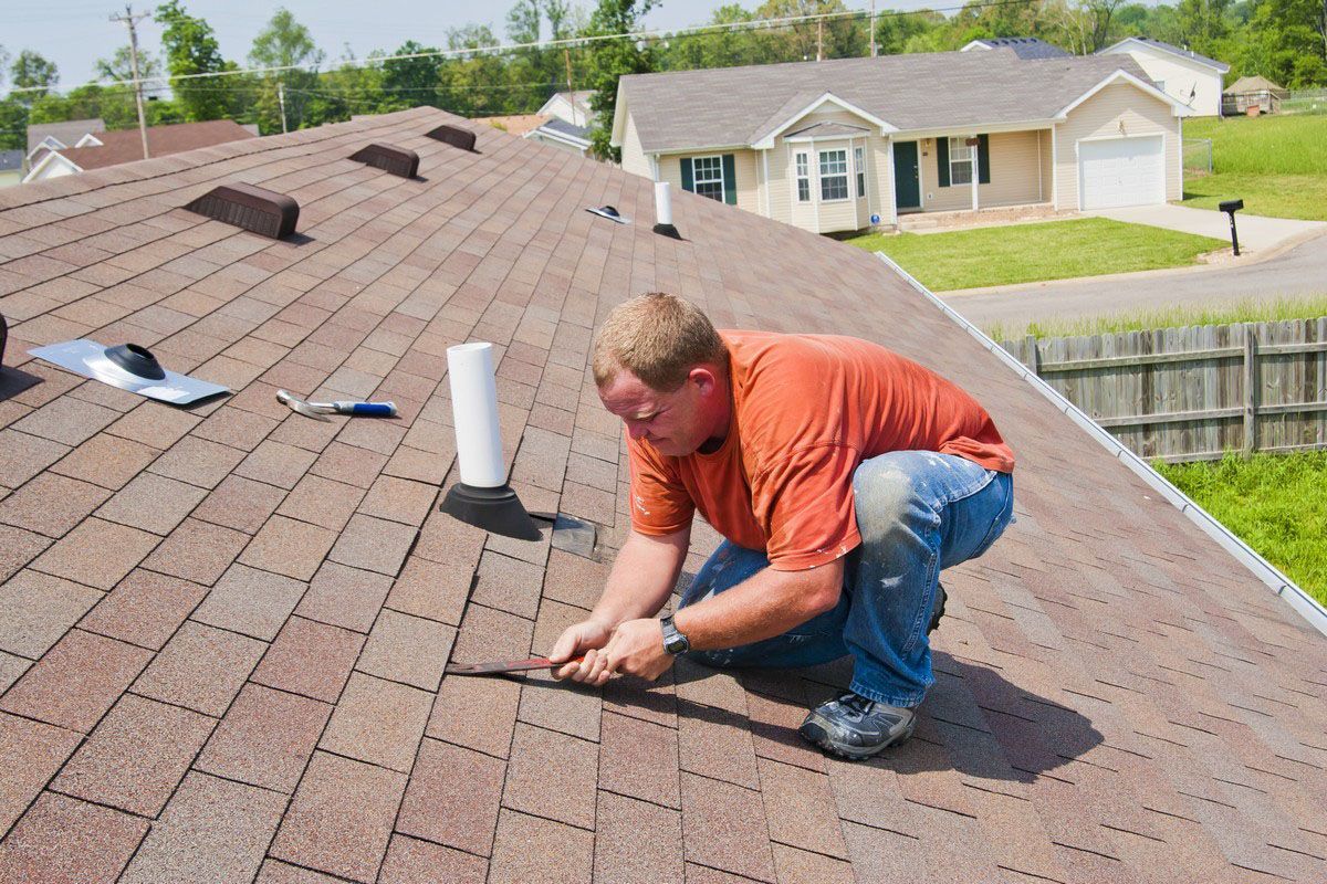 Roofer repairs roof shingles on a house. Brown shingles, orange shirt, blue jeans.