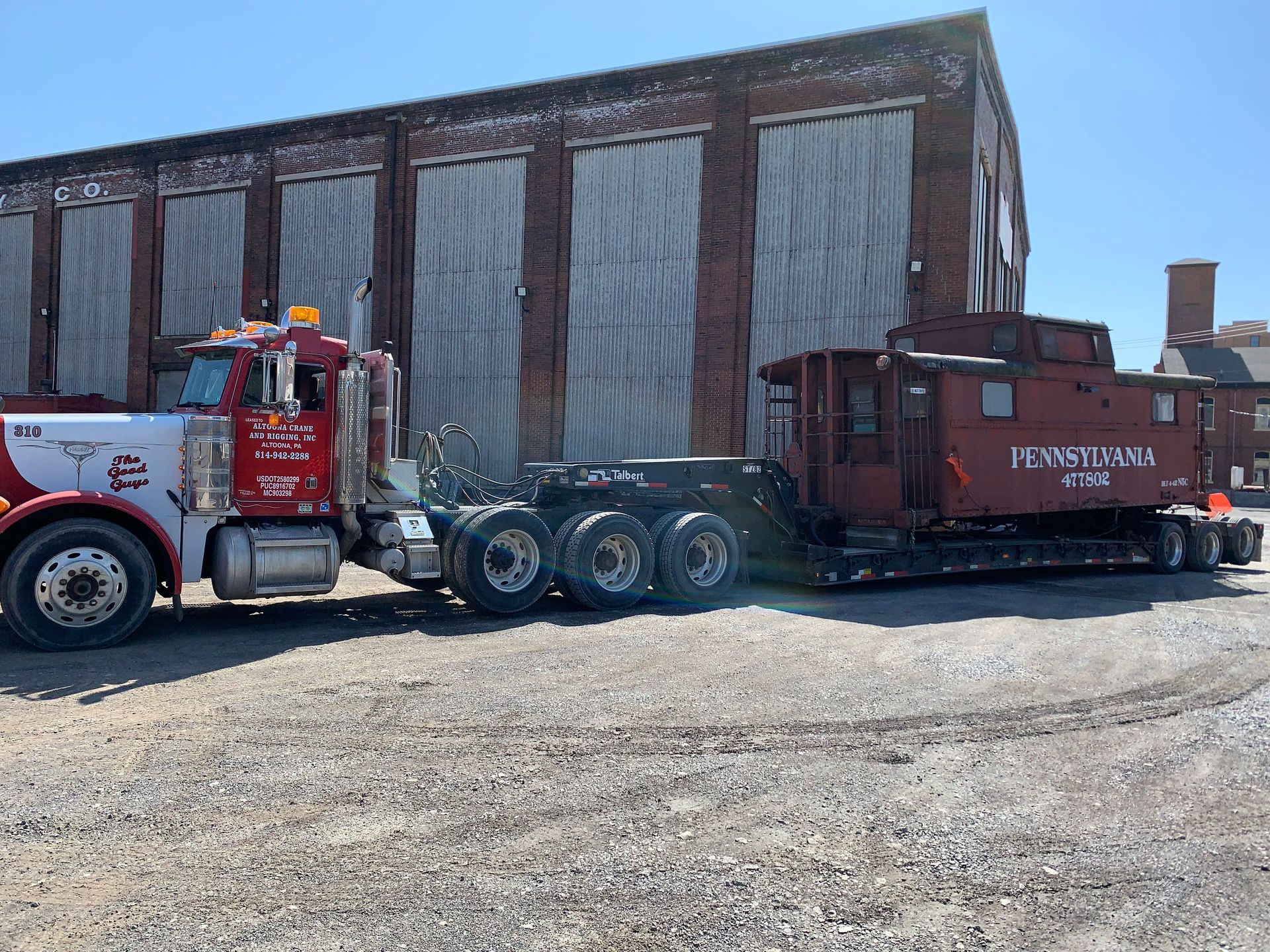 Tow truck hauling a red trailer beside a brick warehouse on a gravel lot