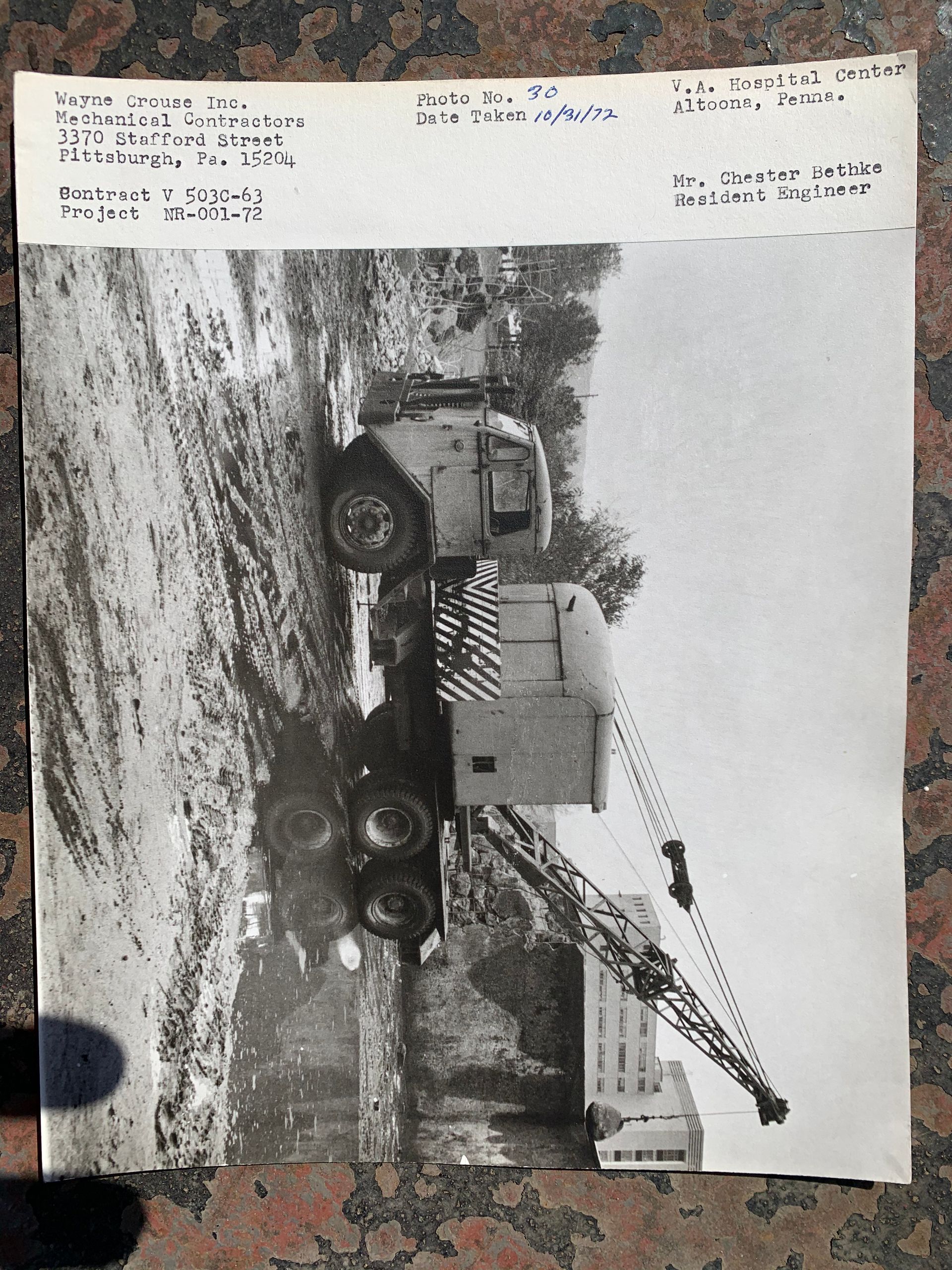 Black-and-white photo of a crane truck lifting a large container in a muddy industrial yard.