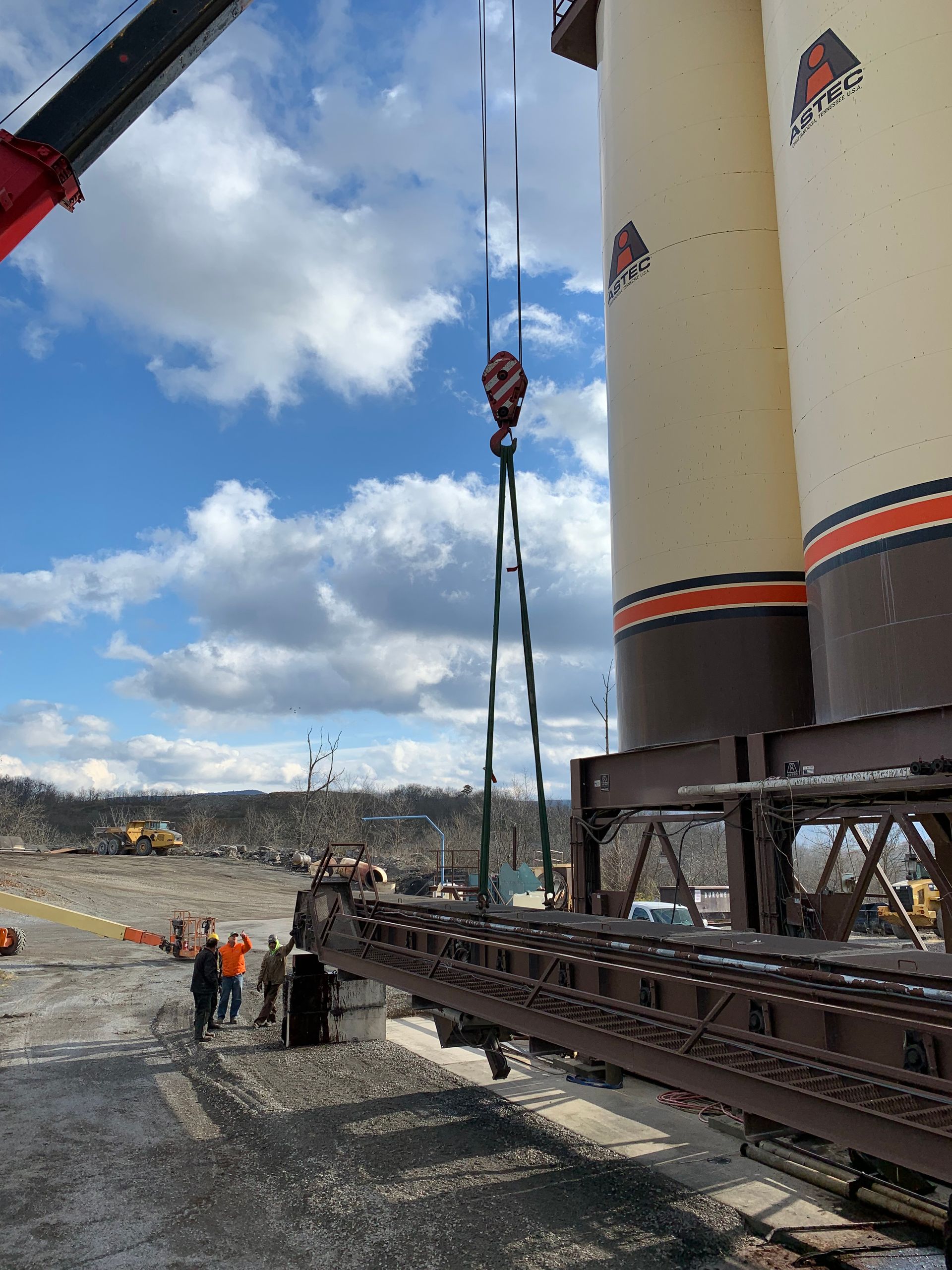 Crane lifting a long steel structure beside a large white industrial tower on a cloudy construction site