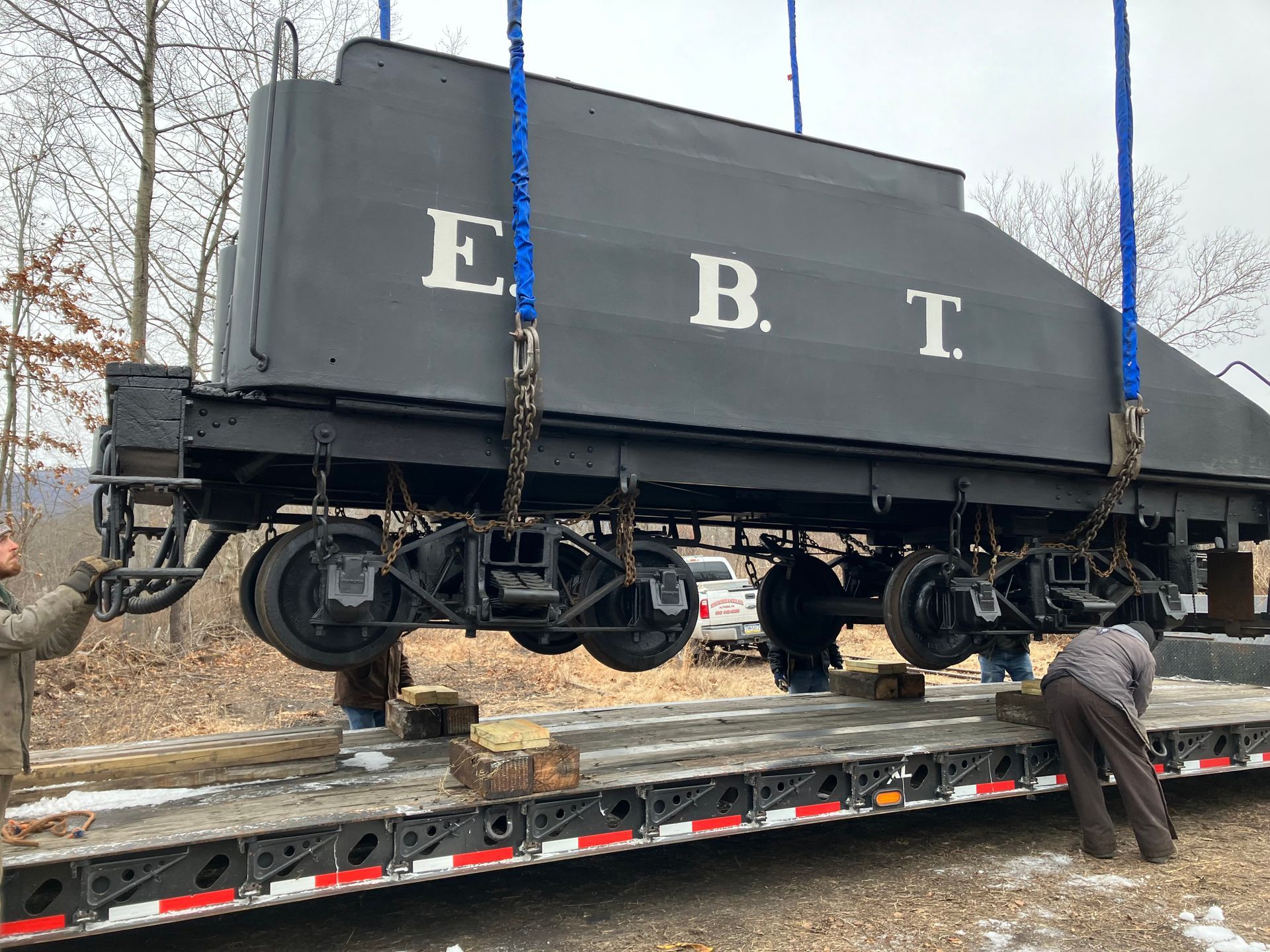 Black EBT railcar being loaded onto a flatbed trailer with chains and workers beside it