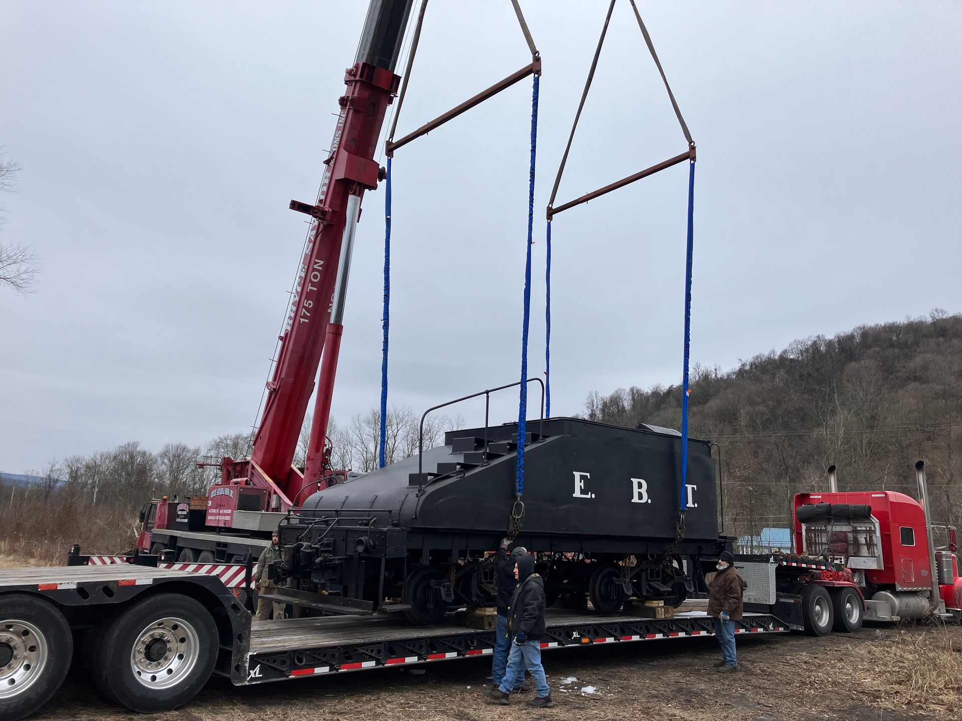 Red crane loading a black rail car onto a flatbed trailer beside a red semi-truck outdoors