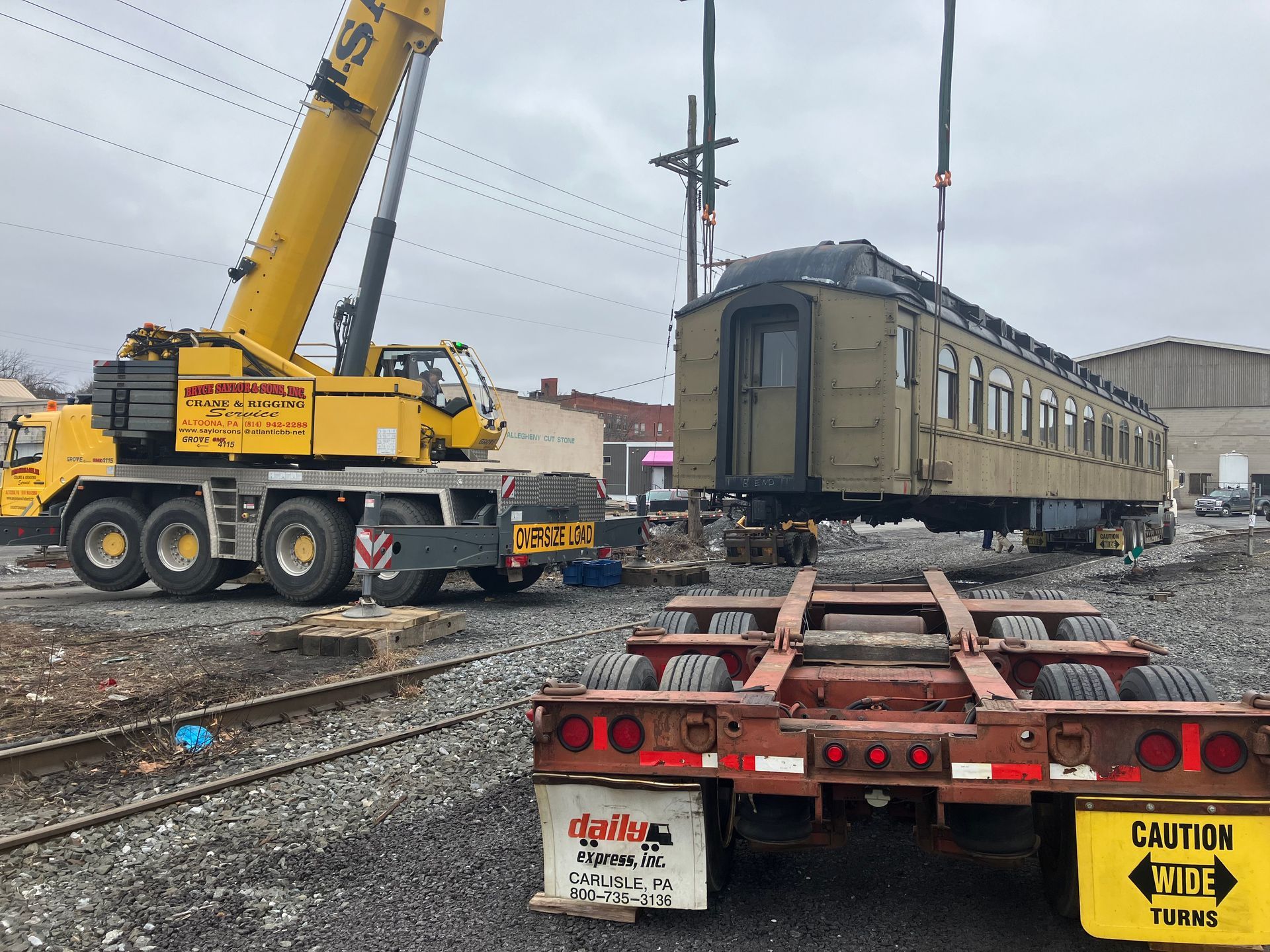 Yellow crane lifting a railcar onto tracks in a rail yard with a flatbed trailer in foreground