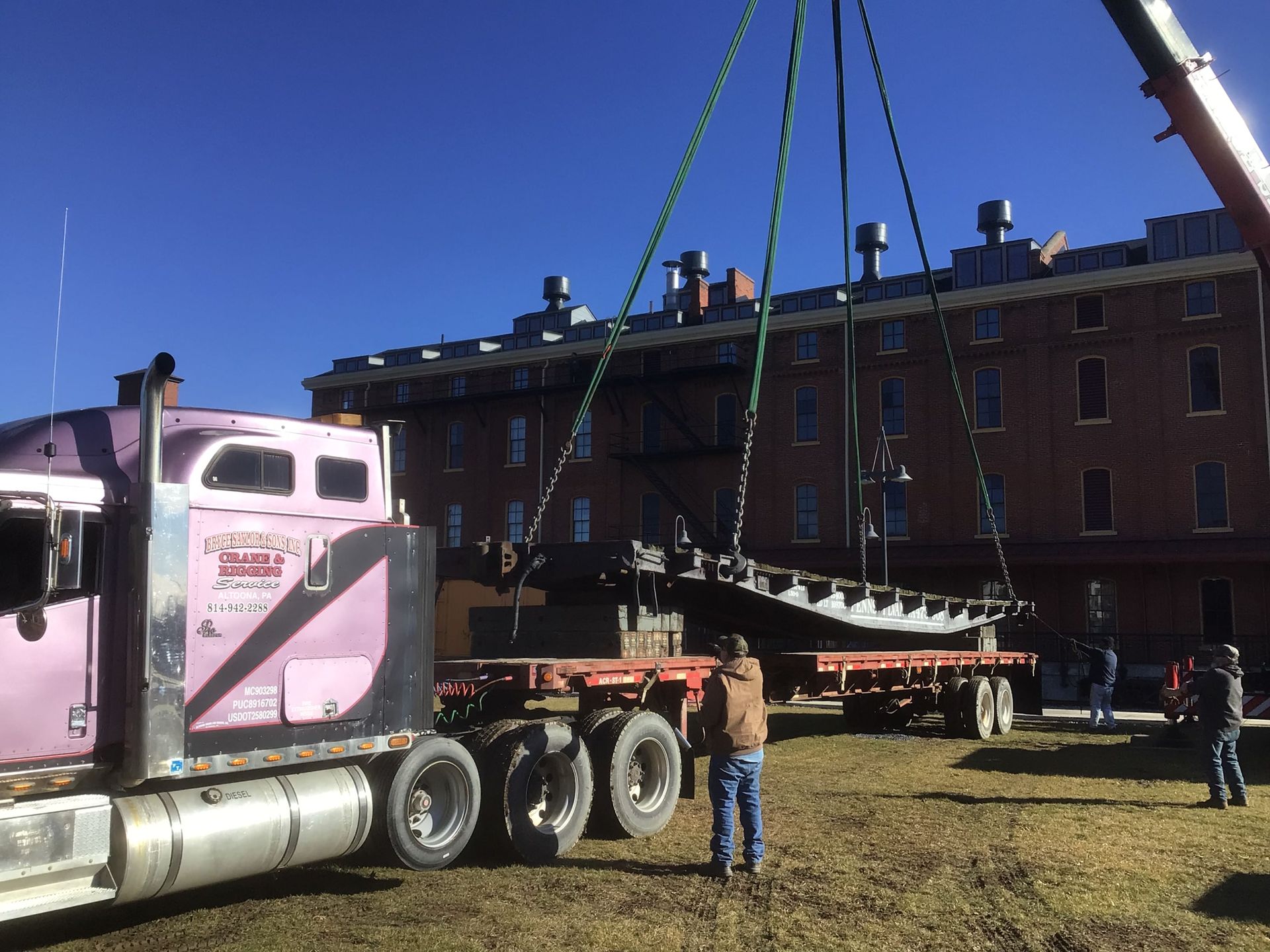 Crane lifting a large load beside a pink semi truck in front of a brick building