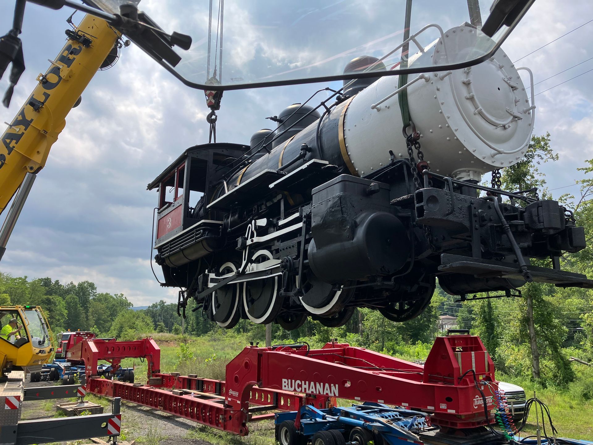 Steam locomotive being lifted by cranes onto a red rail transporter in a grassy field