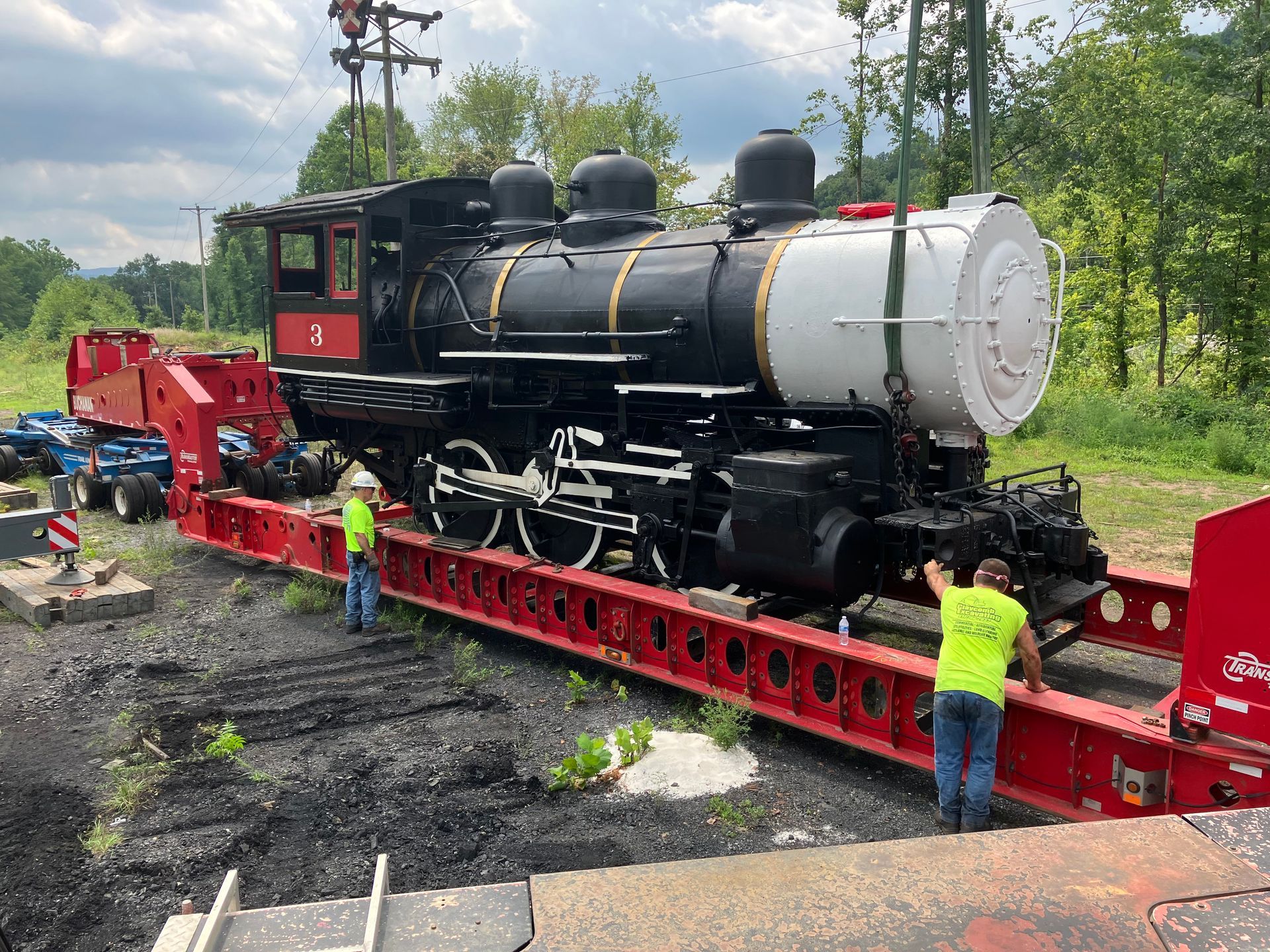 Black steam locomotive on red rail trailer being transported outdoors, with workers nearby
