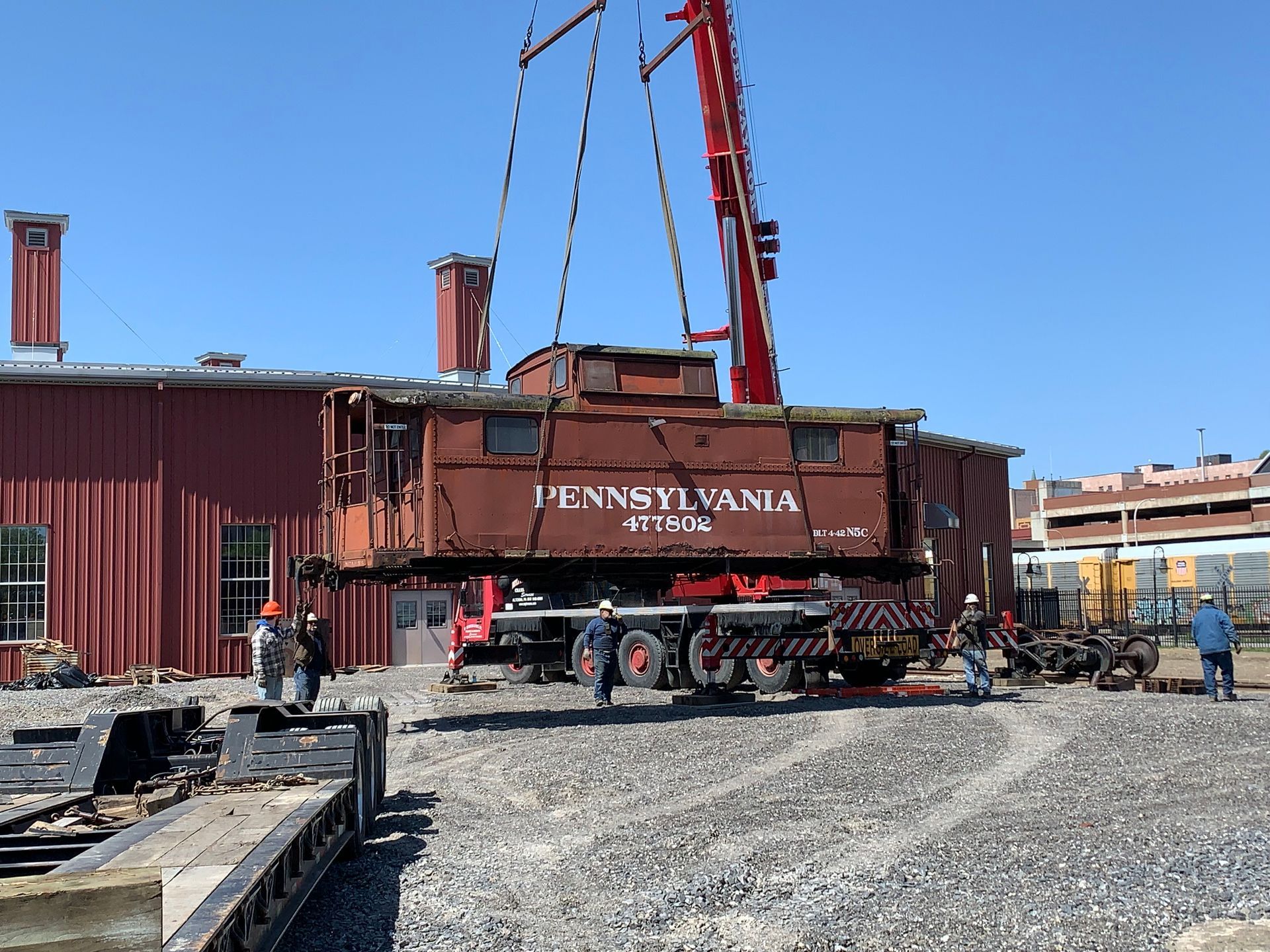 Red railroad caboose being lifted by a crane in an industrial yard under a blue sky