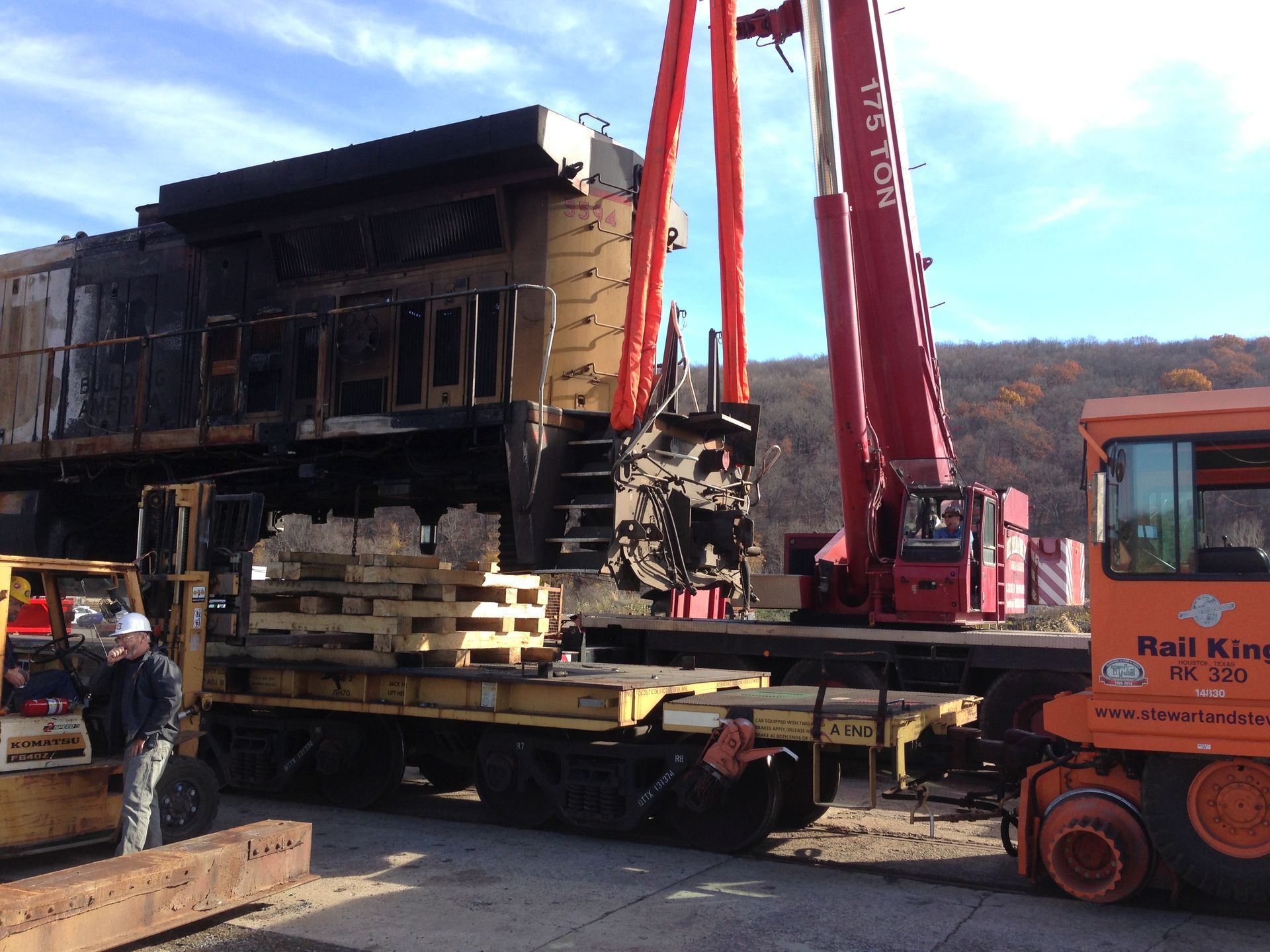 Crane lifting a charred house onto a flatbed truck at a roadside site