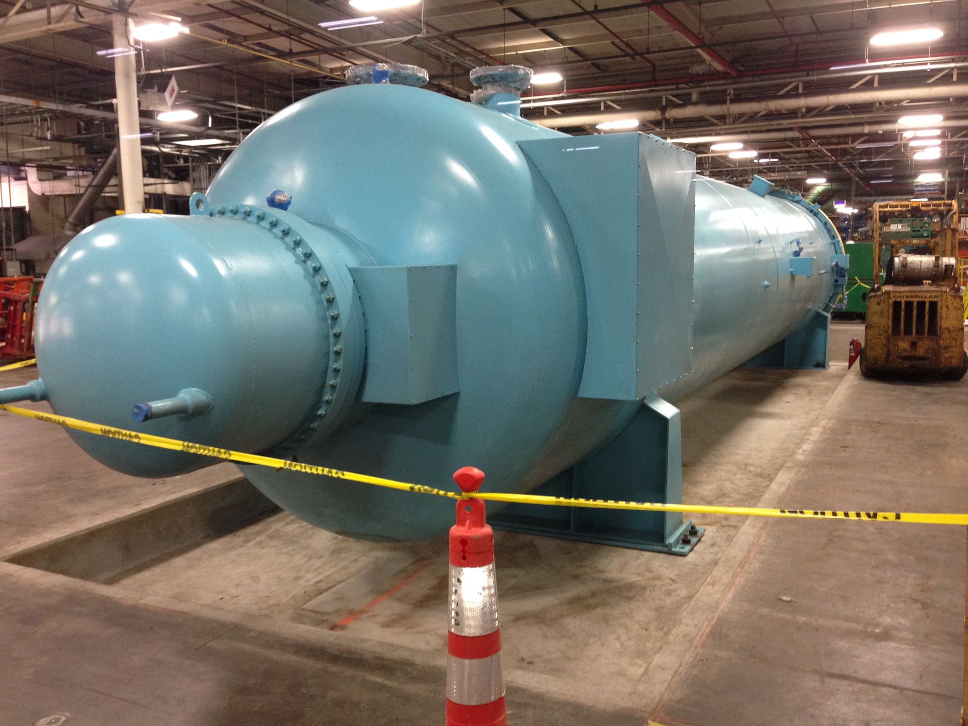 Large light-blue industrial machine or vessel in a factory, behind caution tape and a safety cone