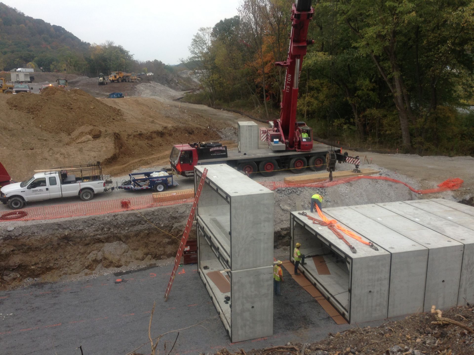 Construction site with crane lifting large concrete blocks beside a road and hillside.