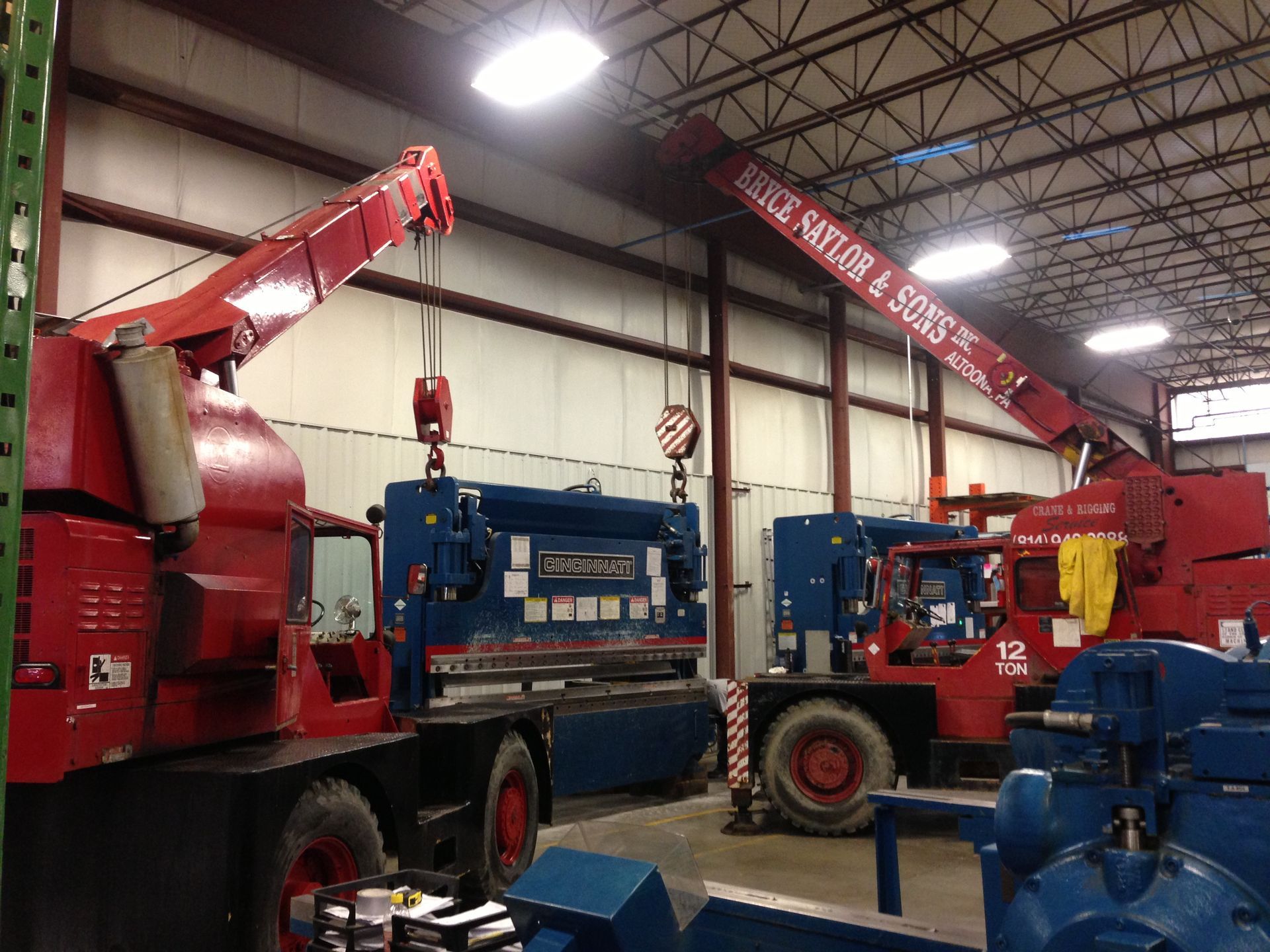 Inside a warehouse, two red cranes lift a blue truck beside a large blue machine.