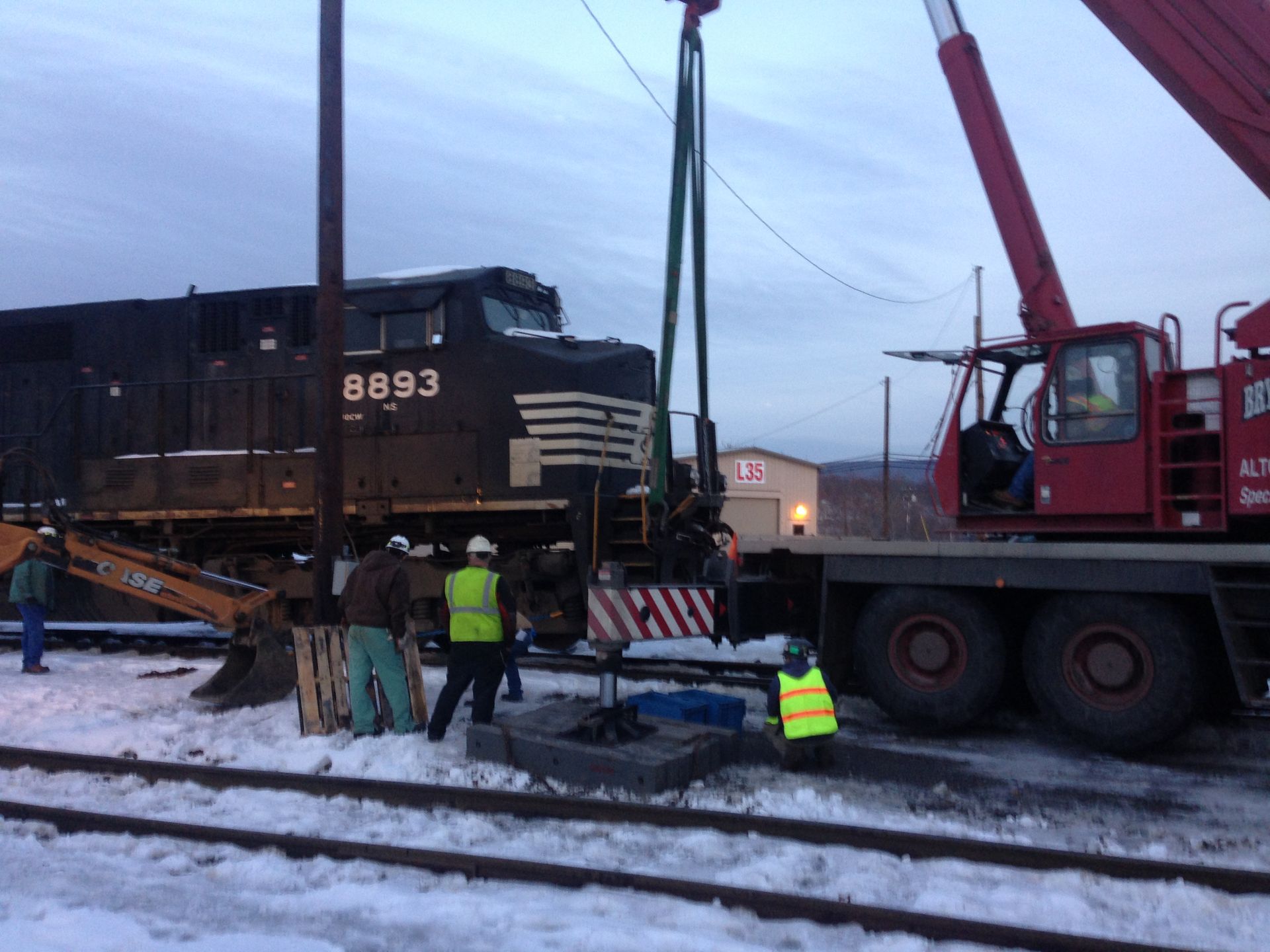 Workers in reflective vests guide a locomotive being lifted by a crane in a snowy rail yard.