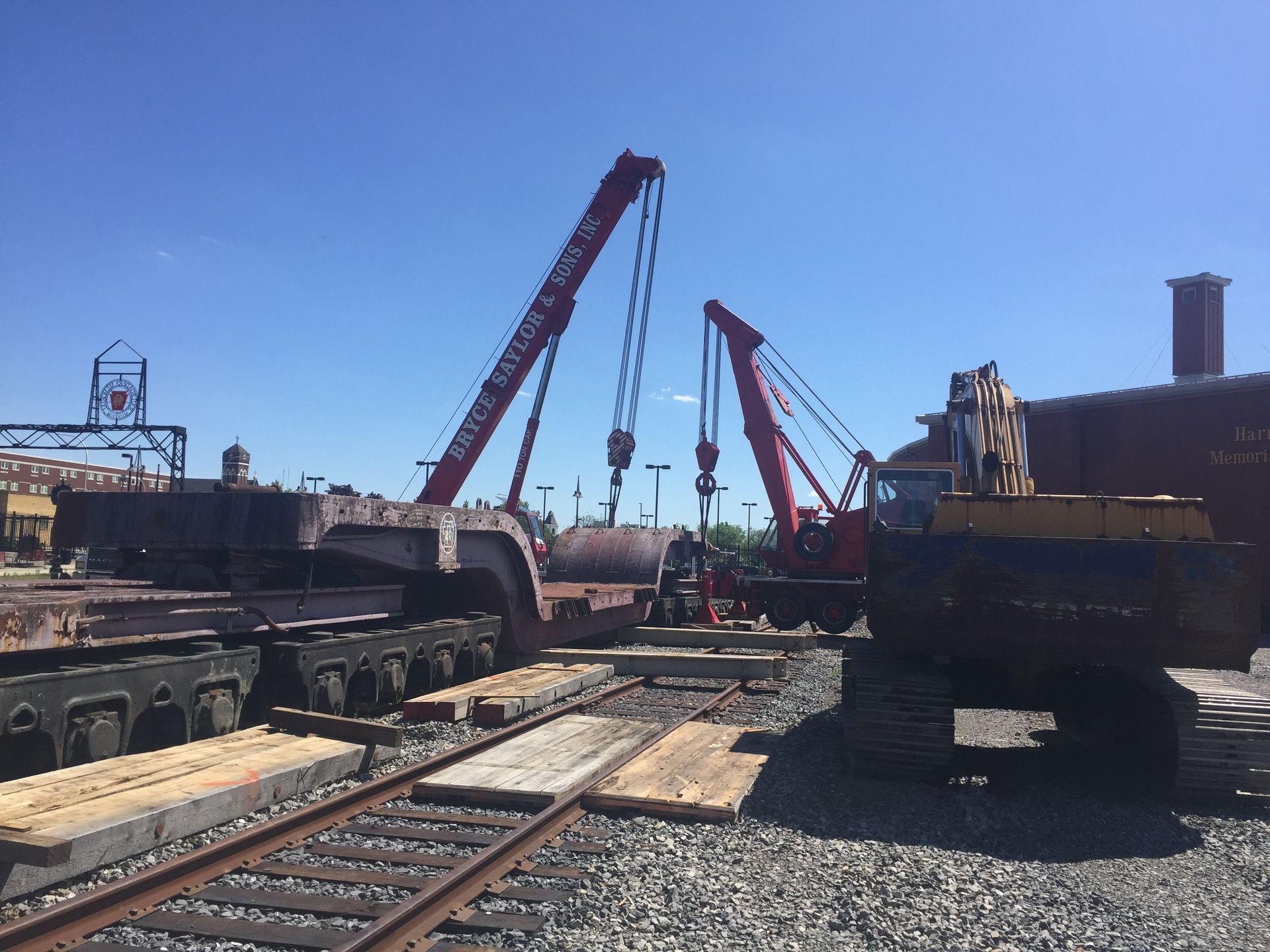 Industrial yard with cranes lifting heavy machinery beside railway tracks under a clear blue sky