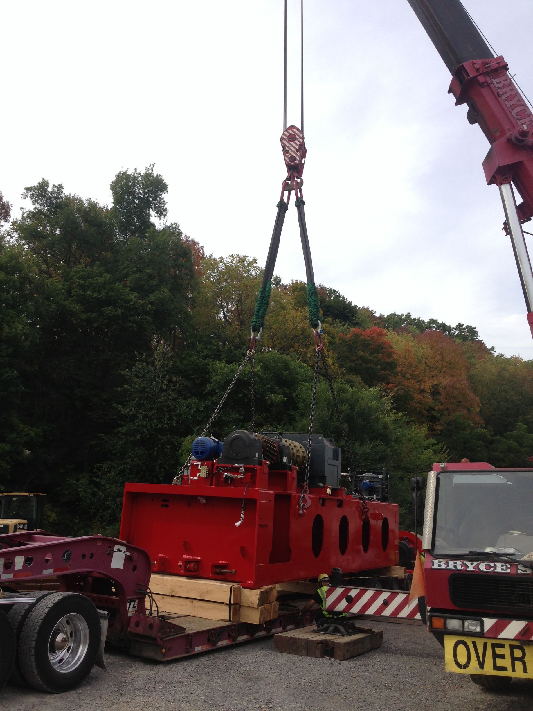 Crane lifting a red industrial machine onto a truck at a roadside work site