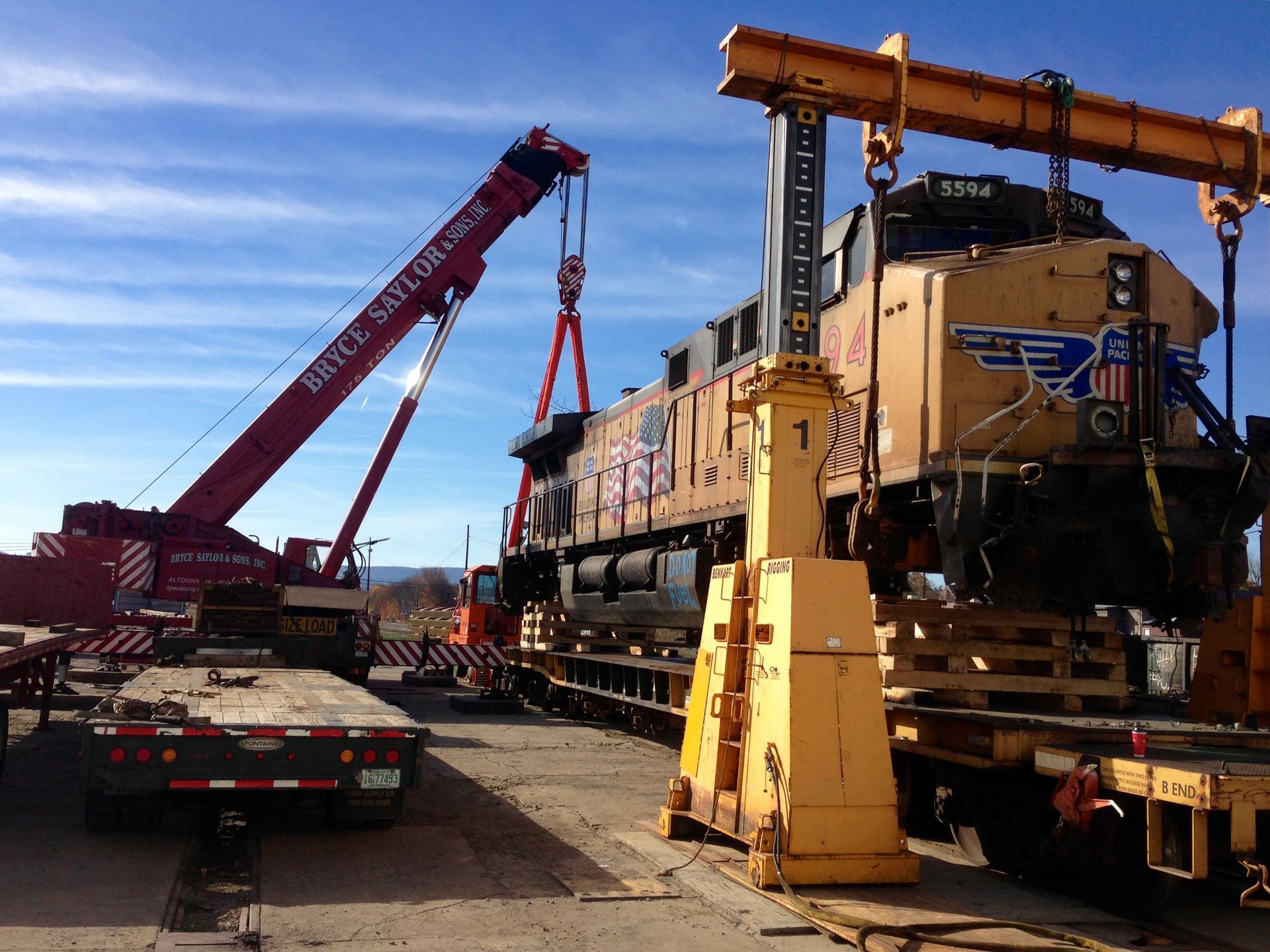Yellow locomotive being lifted by red cranes on a flatbed railcar in a rail yard