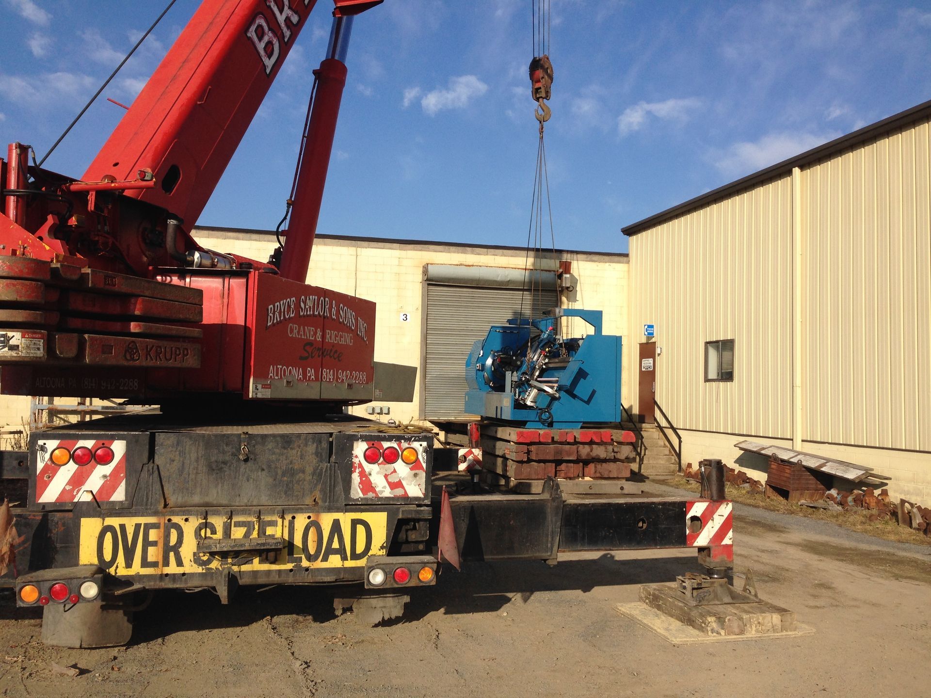 Crane lifting equipment onto a truck beside a warehouse, with an oversize-load sign.