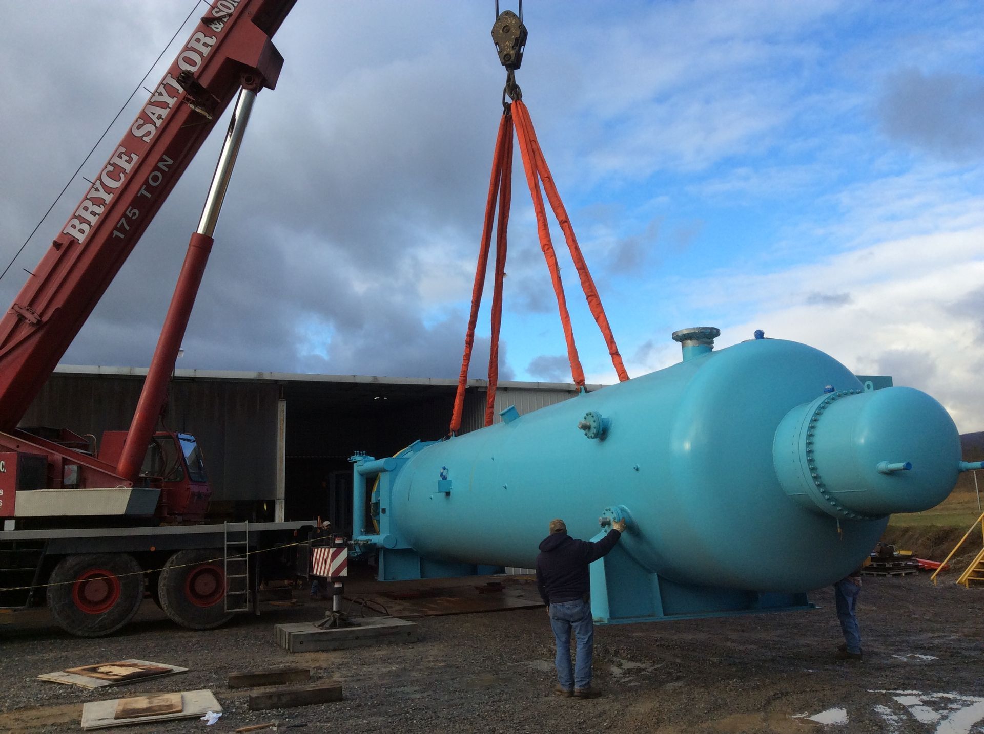 Red crane lifting a large blue industrial tank at a construction site, with a worker nearby.
