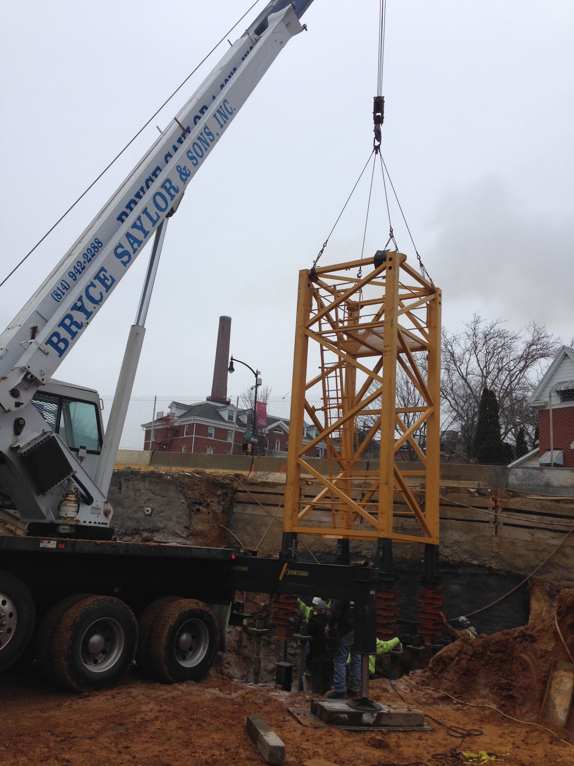 Crane lifting a yellow steel frame at a muddy construction site beneath a cloudy sky
