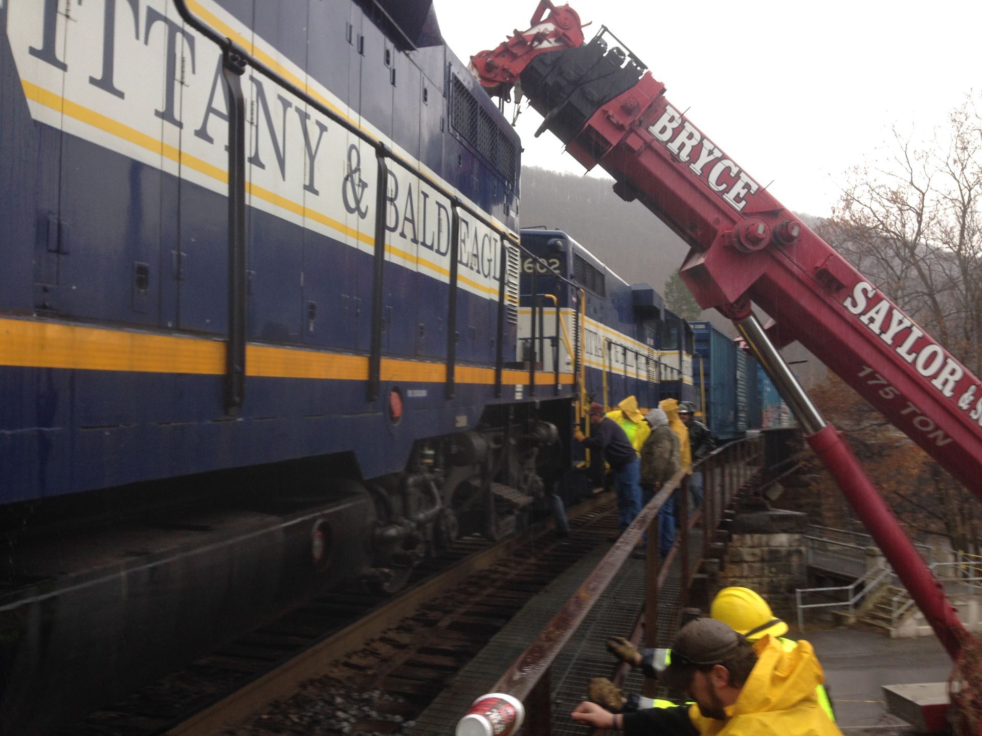 Workers beside derailed train cars, using a red crane to lift a blue and yellow railcar at a trackside scene