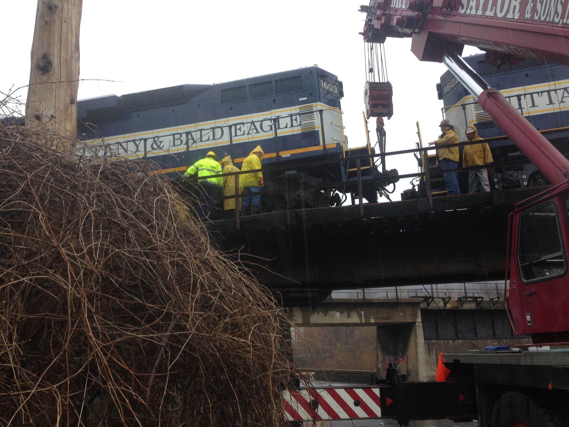 Workers in high-visibility jackets inspect a damaged crane lifting a derailed train car.