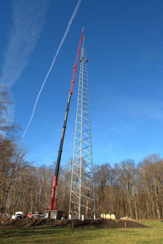 Crane lifting a tall metal tower against a clear blue sky in a wooded field